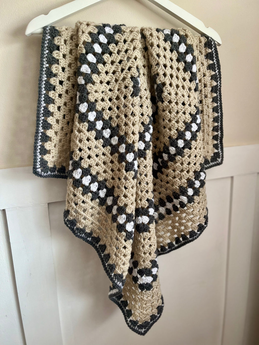 A handmade crochet baby blanket with a grey and beige pattern, hanging from a white wooden hanger against a white paneled wall.