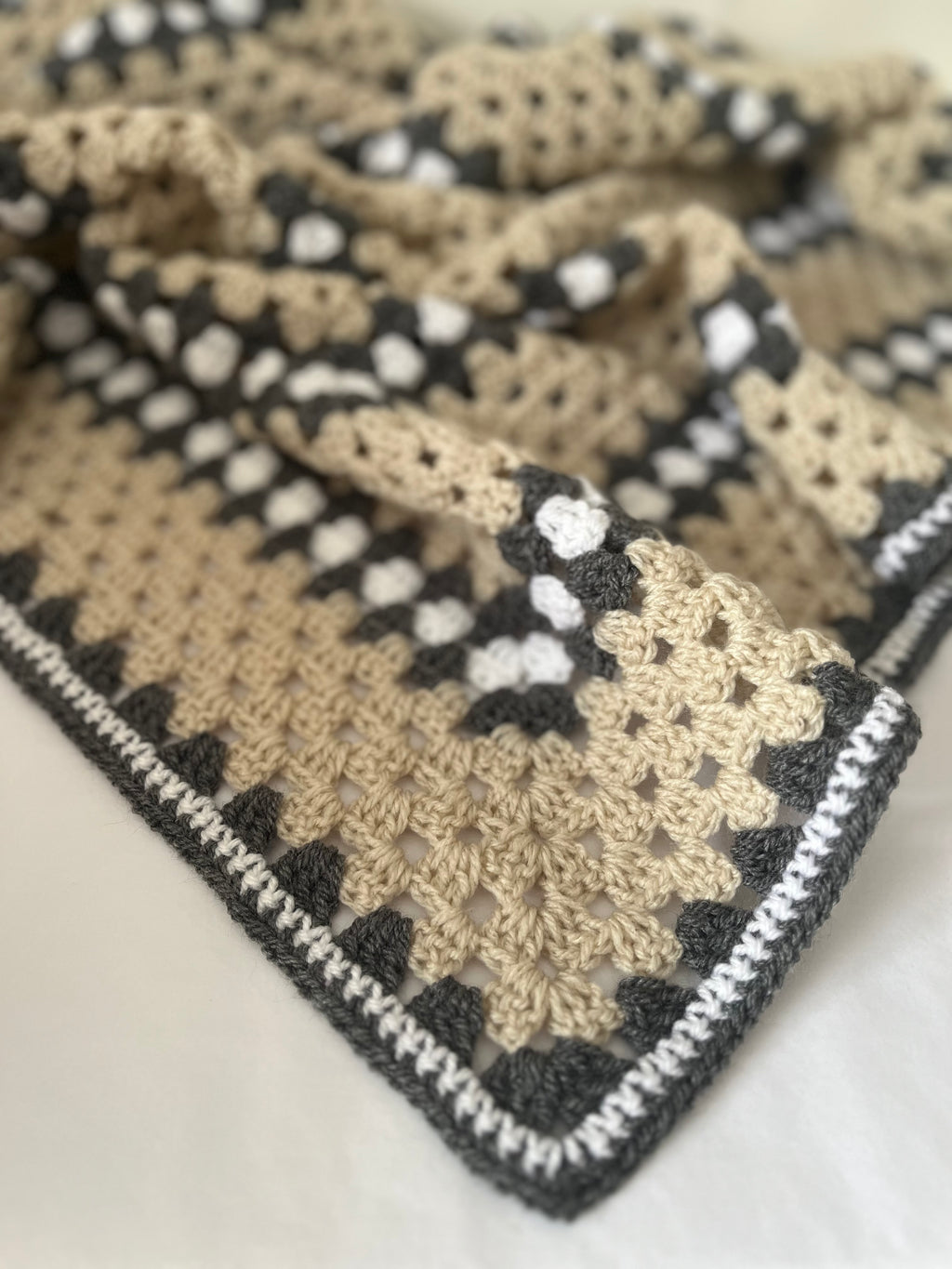 A close-up of a crocheted blanket with a geometric pattern in beige, dark gray, and white, laid on a light neutral surface.