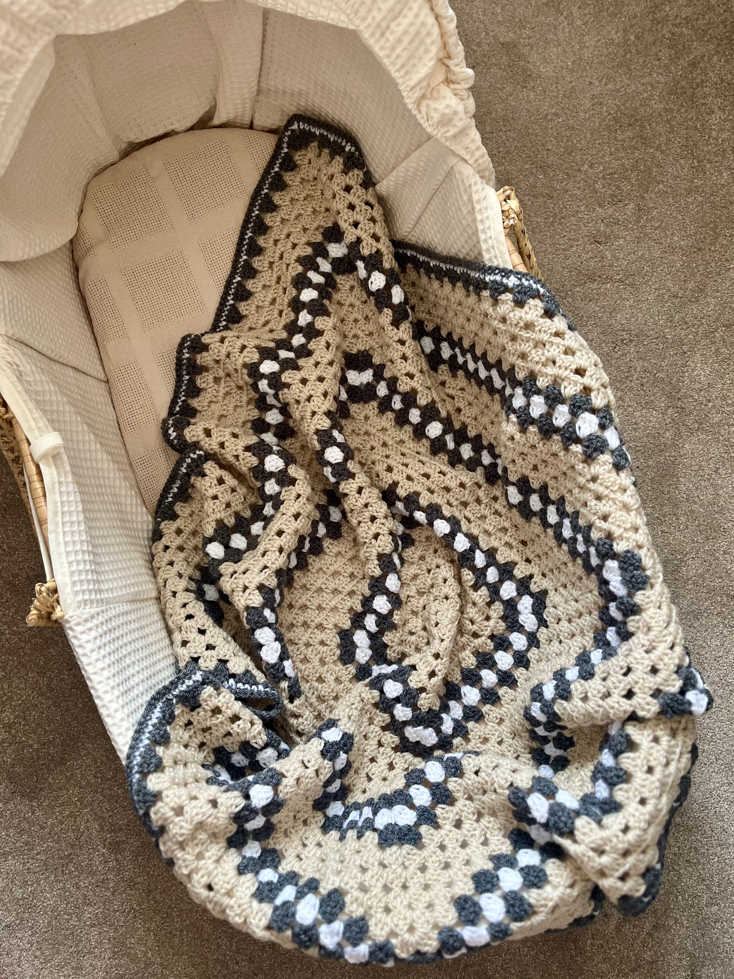 A handmade crochet baby blanket with a beige base and dark grey and white geometric patterns, draped over a woven bassinet.