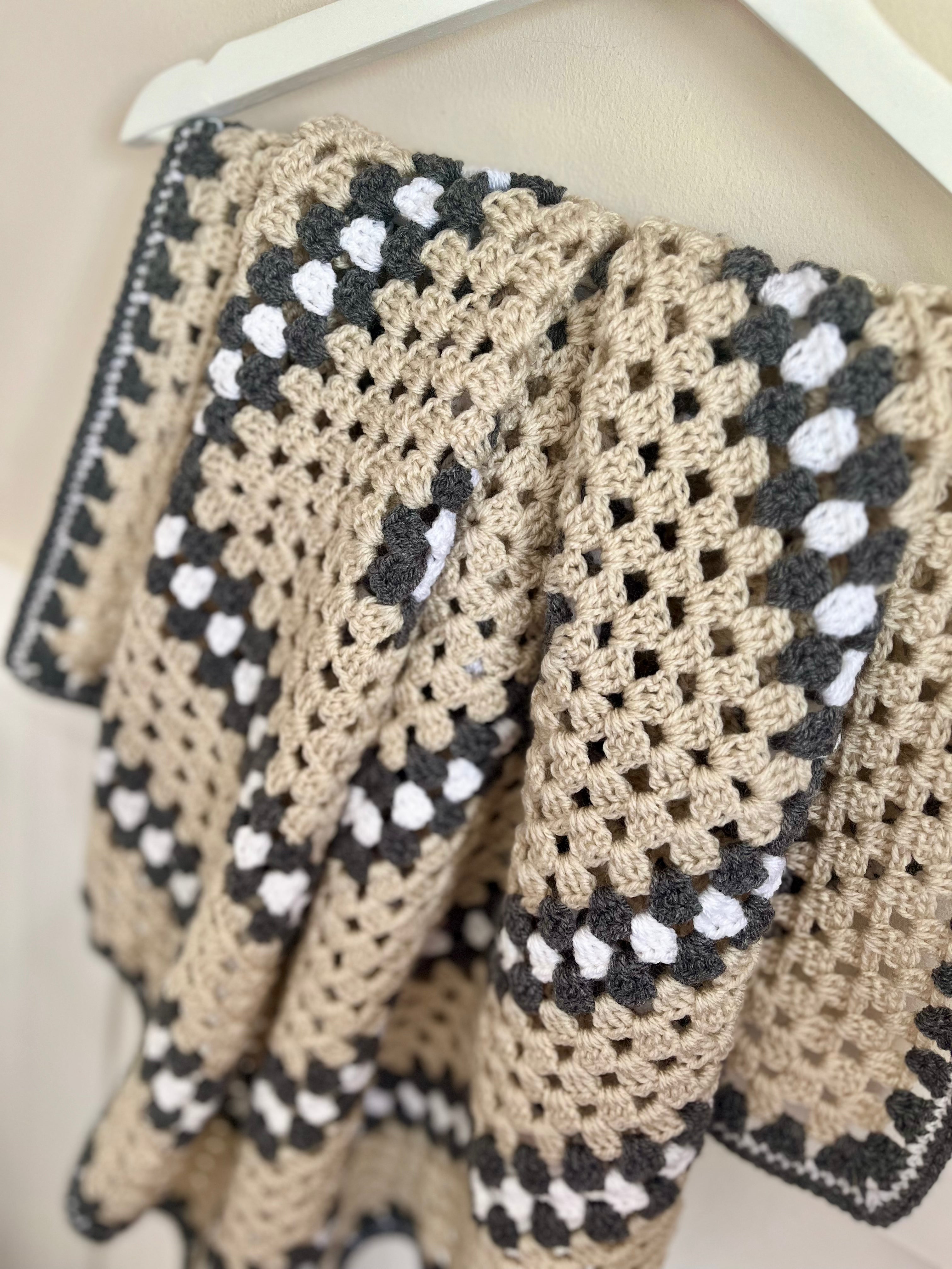 A close-up of a crocheted blanket draped over a white hanger, featuring a beige base with grey and white granny square patterns.