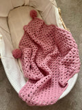 A dusty pink crocheted baby blanket with two pom-poms, draped over a white bassinet.