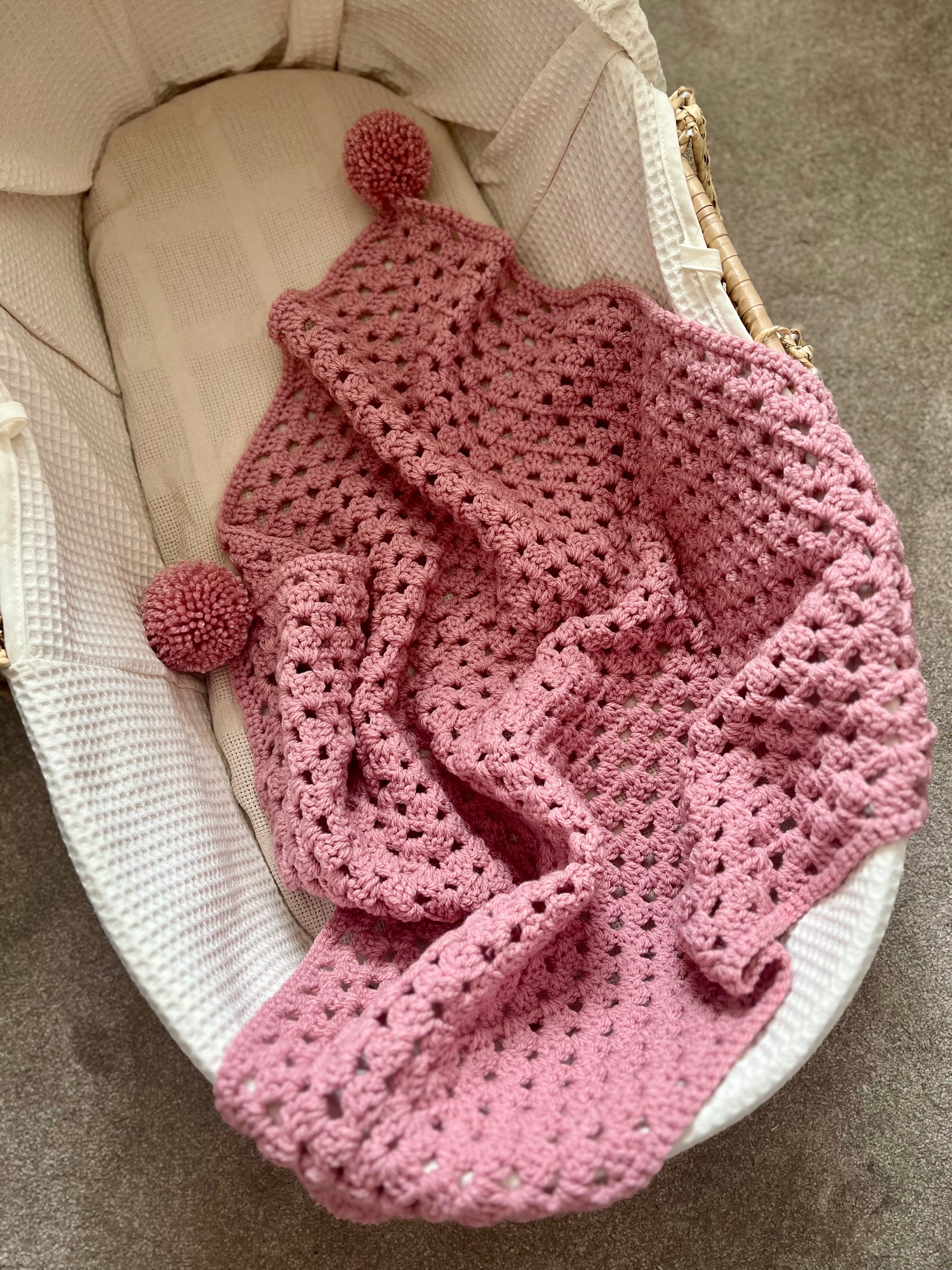 A dusty pink crocheted baby blanket with two pom-poms, draped over a white bassinet.