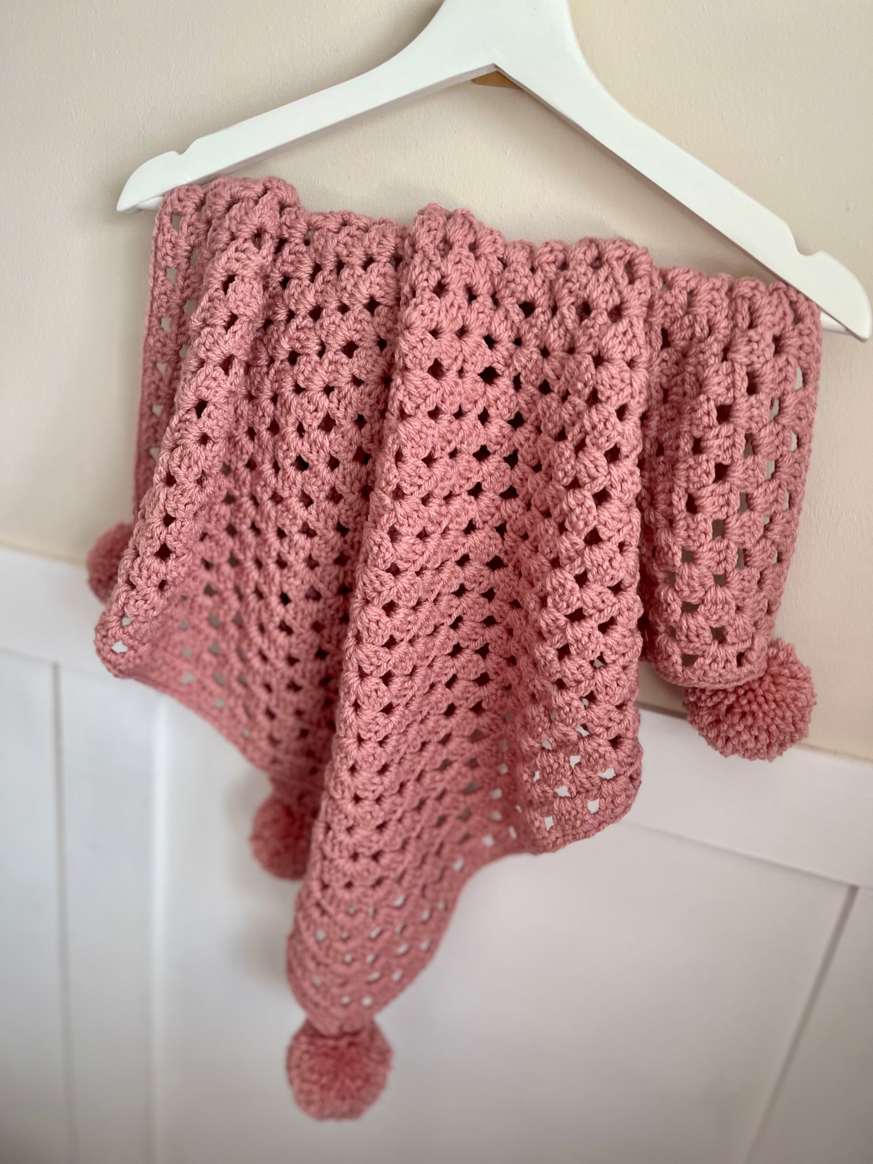 A dusty pink crocheted baby shawl with pom-pom ends, draped over a white plastic hanger against a light beige wall and white paneling.