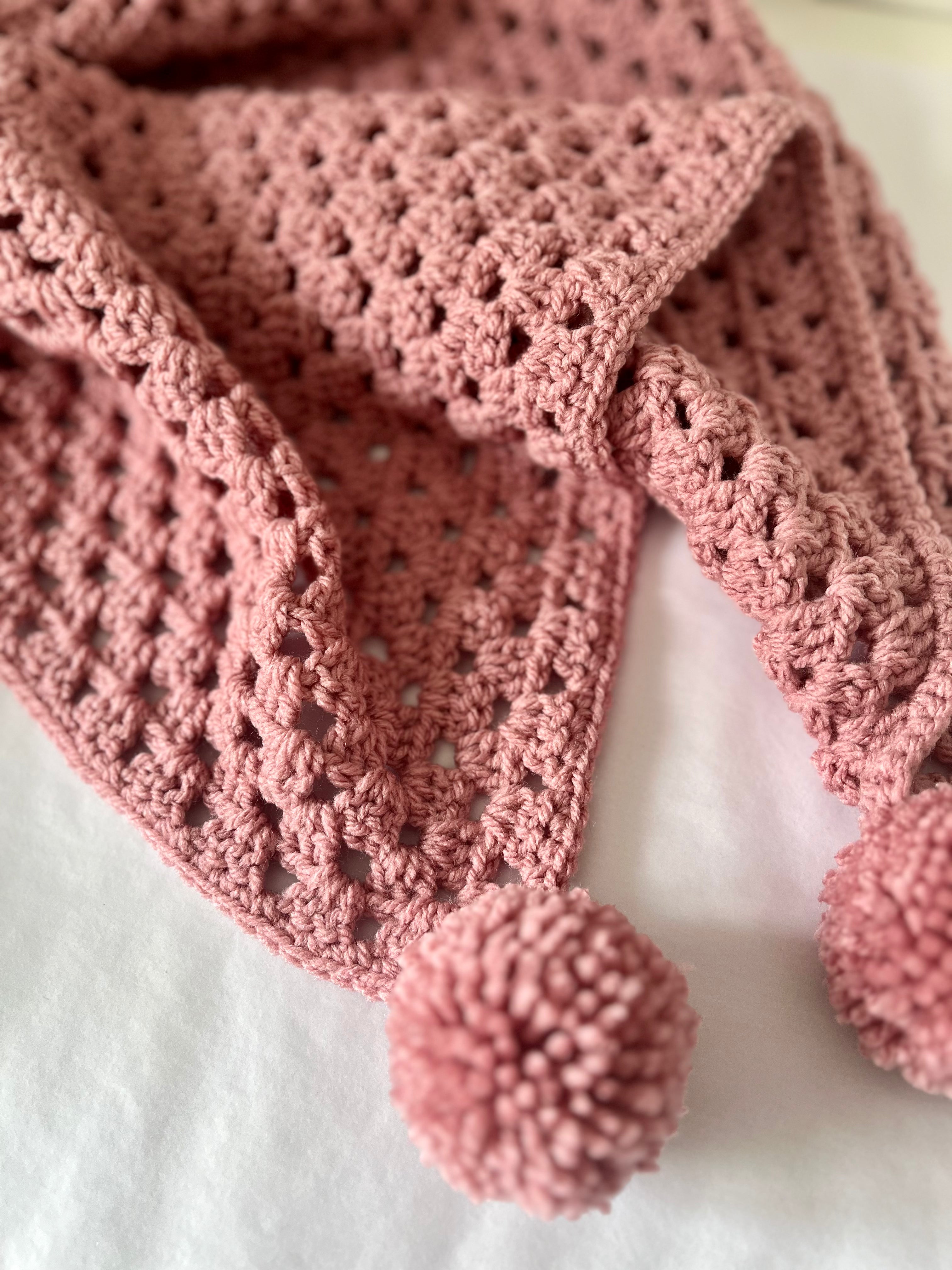A dusty pink crocheted baby blanket with a textured openwork pattern and two matching pom-poms, draped on a white fabric background.