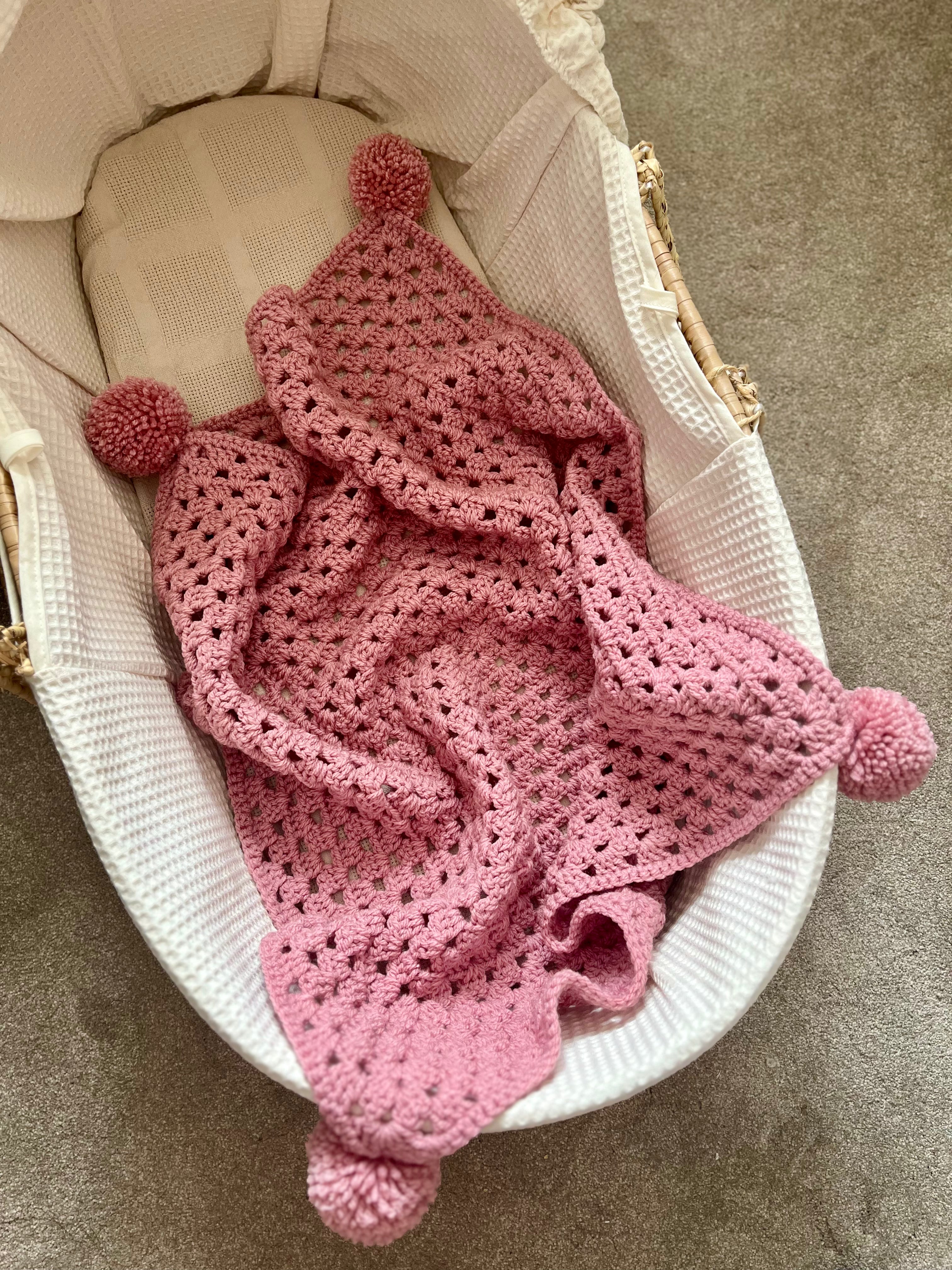 A pink crocheted baby blanket with pom-pom ends draped over a white waffle-weave bassinet on a carpeted floor.