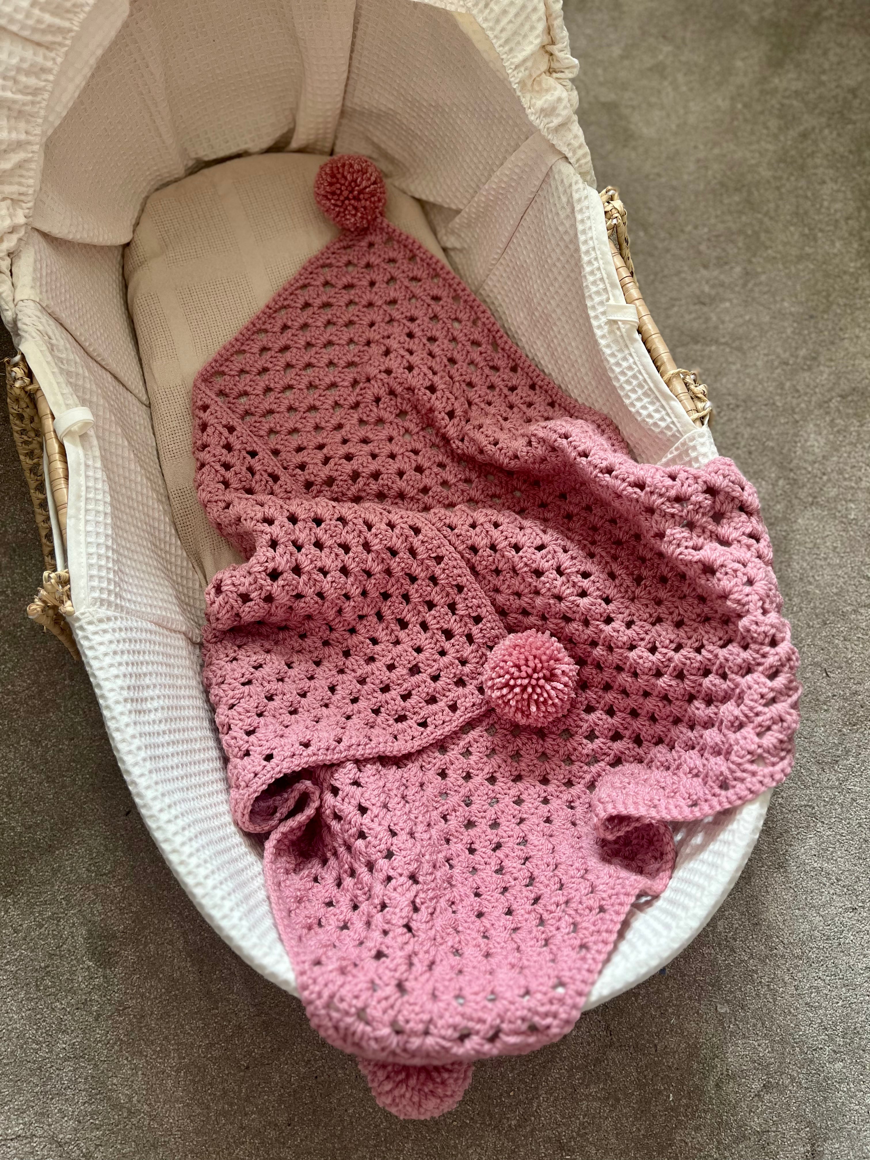 A pink crocheted baby blanket with matching pom-pom accents is draped inside a white waffle-weave bassinet on a gray carpeted floor.