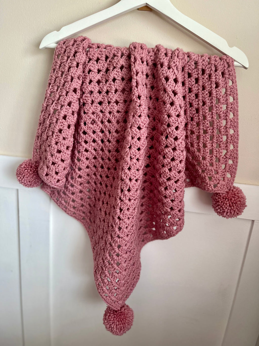 A dusty pink crocheted baby blanket with pom-pom details, hanging from a white hanger against a beige wall and white paneling.