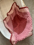 A handmade crocheted baby blanket with a heart design in shades of pink and red, draped over a white woven chair on a gray carpet.