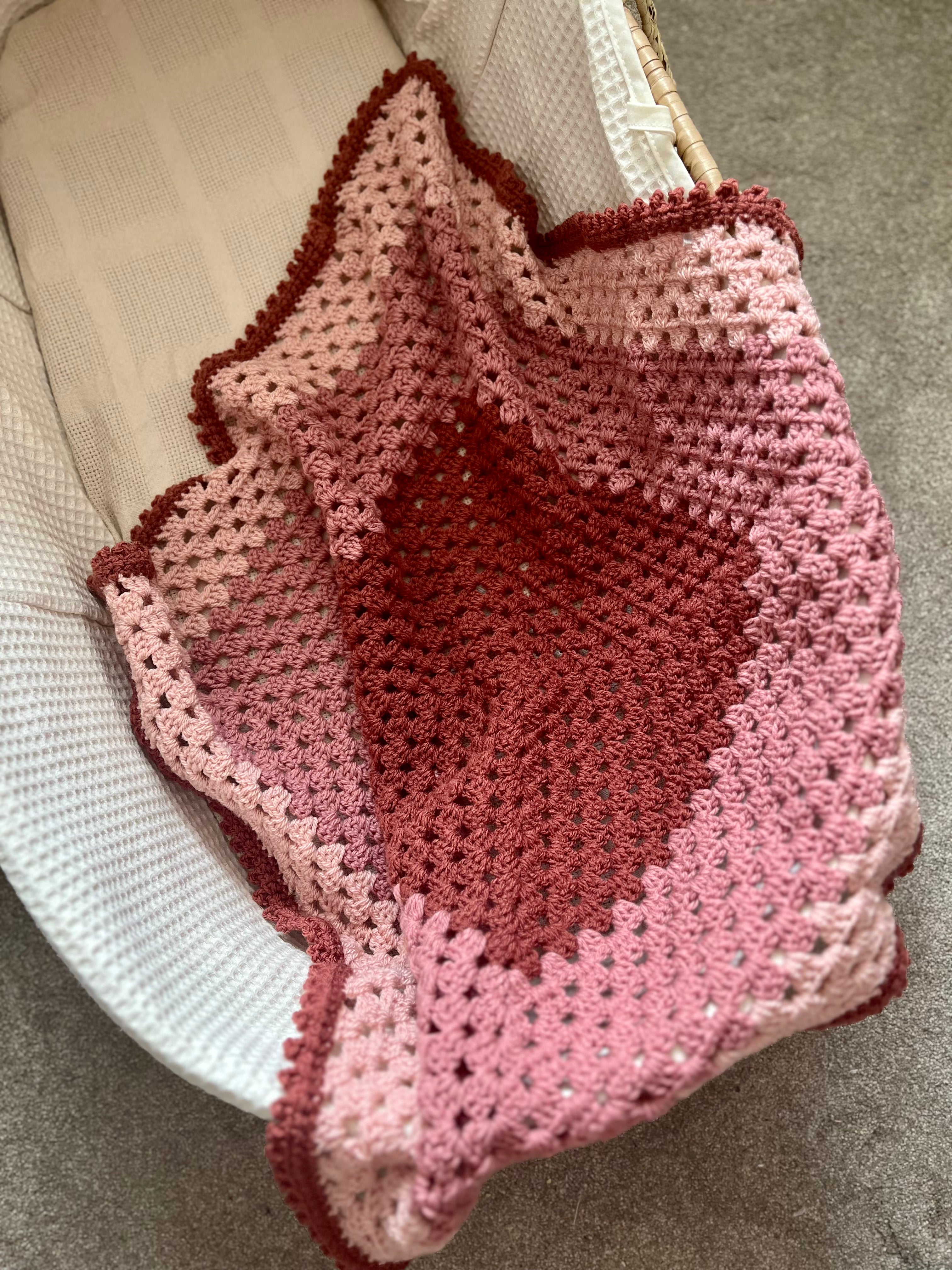 A handmade crocheted baby blanket with a heart design in shades of pink and red, draped over a white woven chair on a gray carpet.