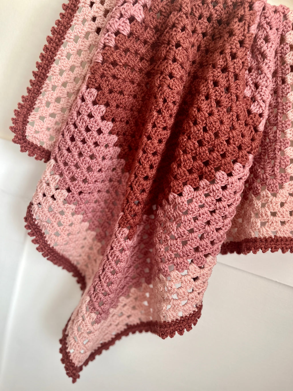 A handmade crocheted baby blanket draped over a white surface, featuring a color gradient from dusty rose pink to terracotta red, with a matching pink and red scalloped border.