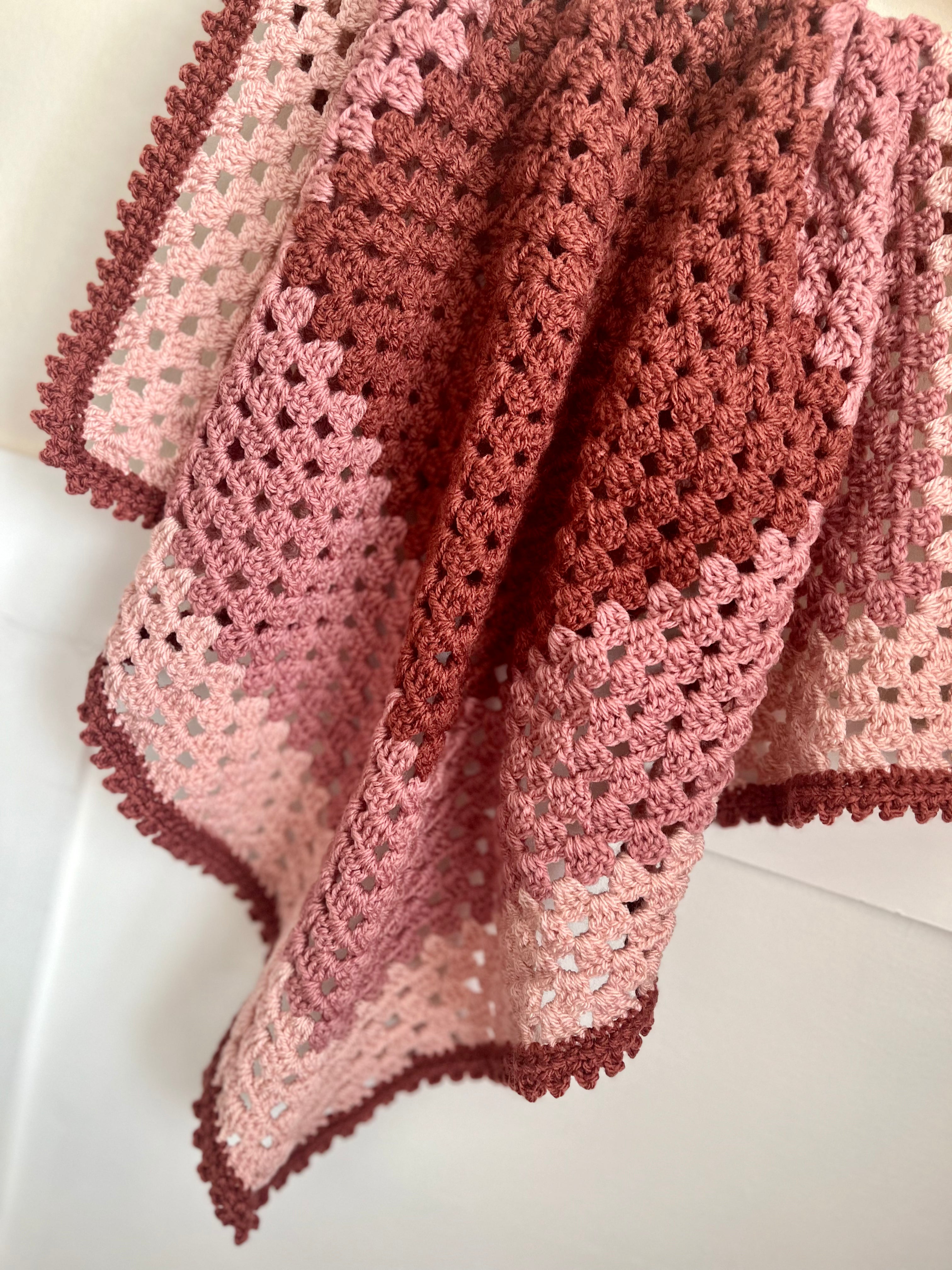A handmade crocheted baby blanket draped over a white surface, featuring a color gradient from dusty rose pink to terracotta red, with a matching pink and red scalloped border.