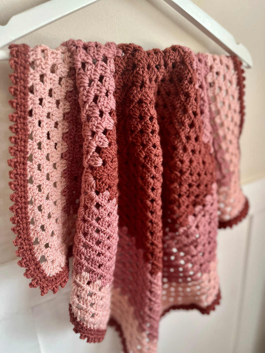 A handmade crocheted blanket with a pink gradient pattern, hanging from a white hanger against a light beige wall.