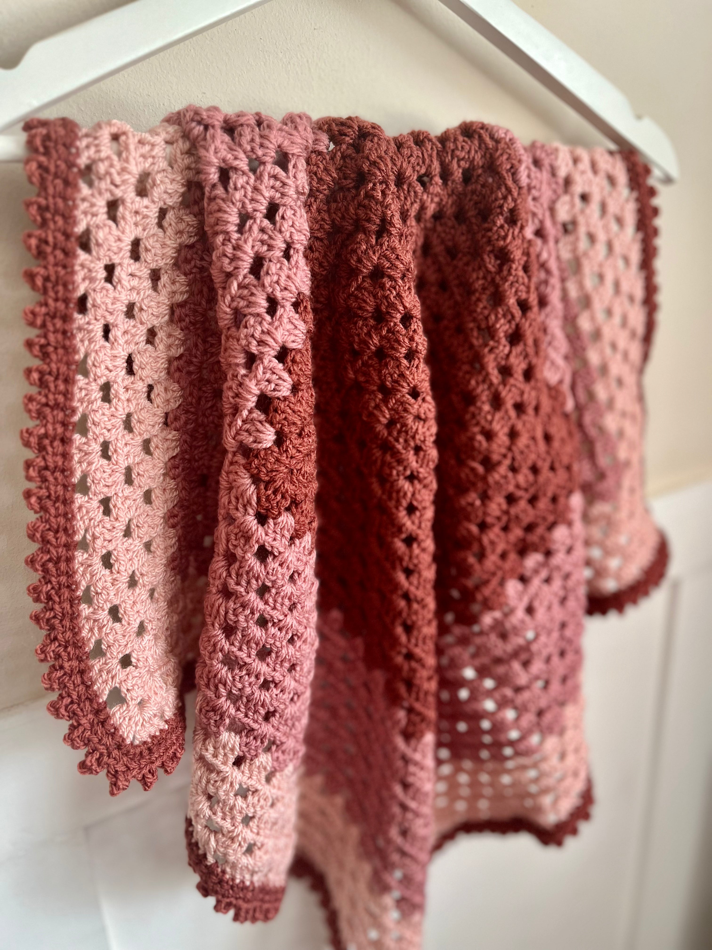 A handmade crocheted blanket with a pink gradient pattern, hanging from a white hanger against a light beige wall.