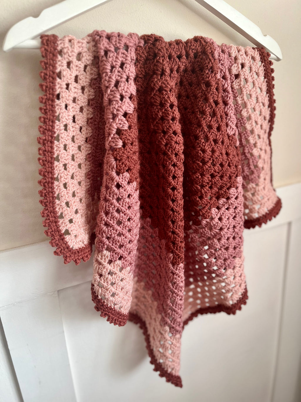 A handmade crocheted shawl or throw blanket in a dusty rose and terracotta gradient, draped over a white hanger against a white paneled wall.