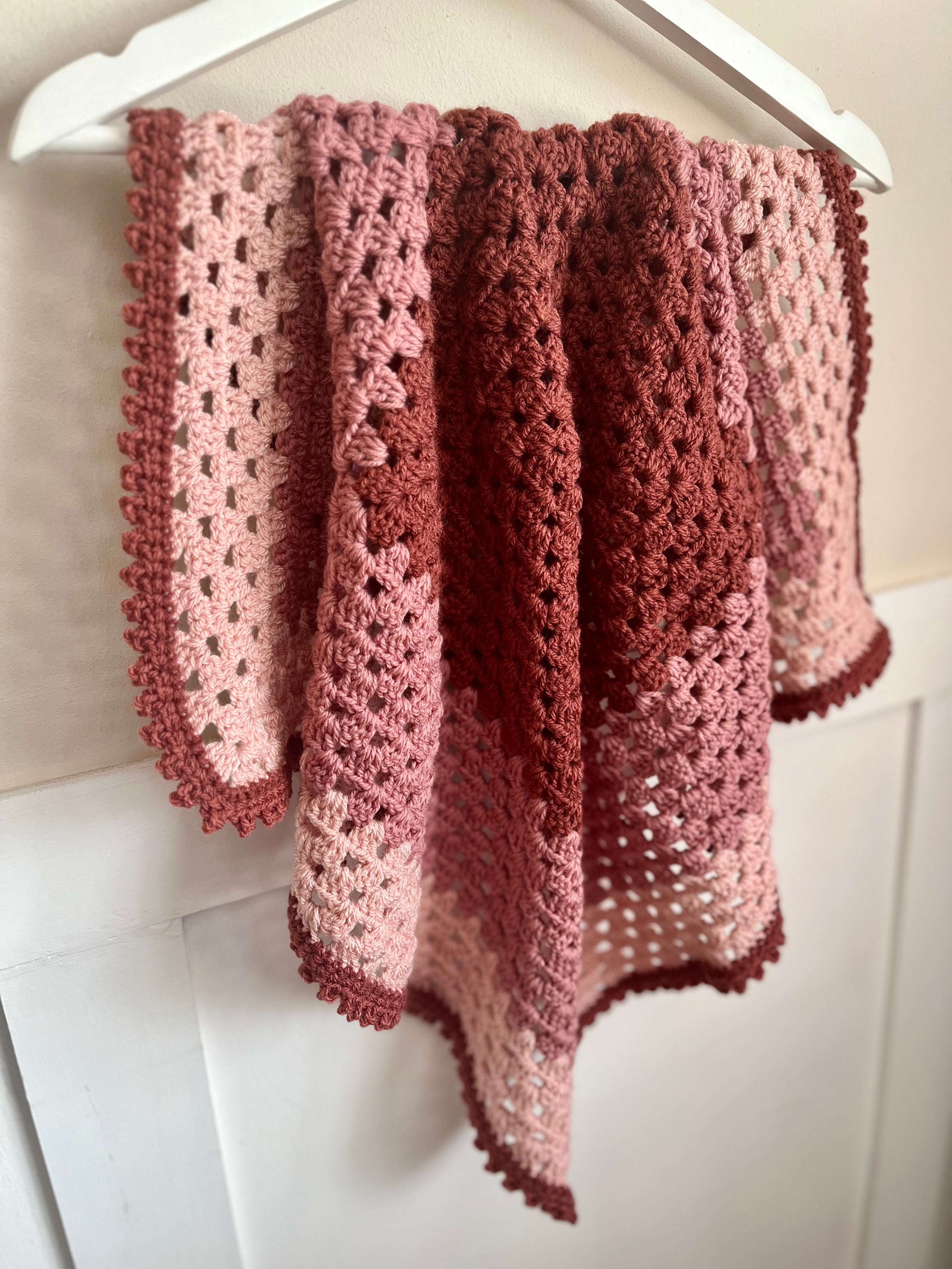 A handmade crocheted shawl or throw blanket in a dusty rose and terracotta gradient, draped over a white hanger against a white paneled wall.