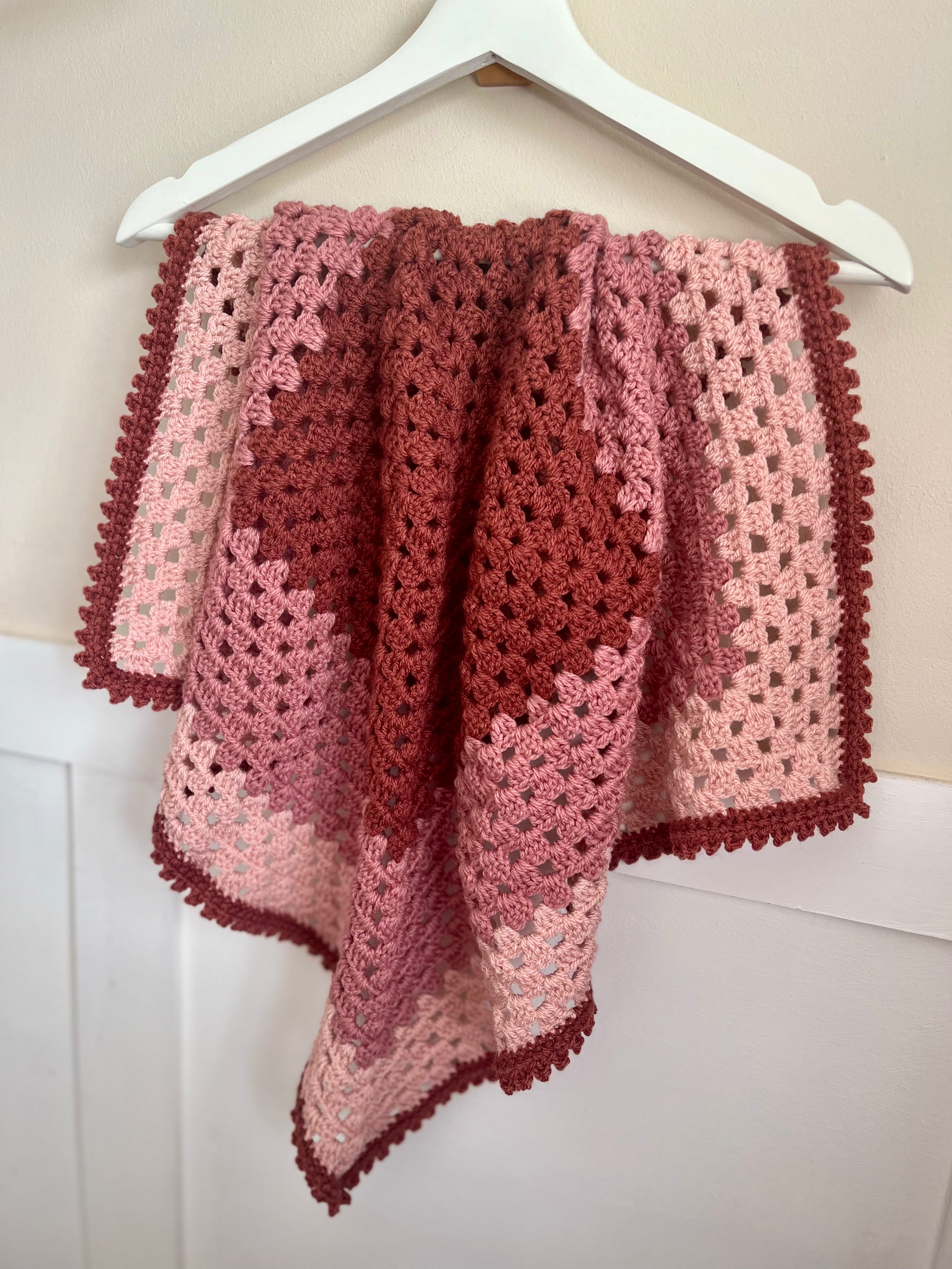 A handmade crocheted baby blanket with a patchwork design in shades of dusty rose, terracotta, and light pink, hanging from a white plastic hanger against a white paneled wall.