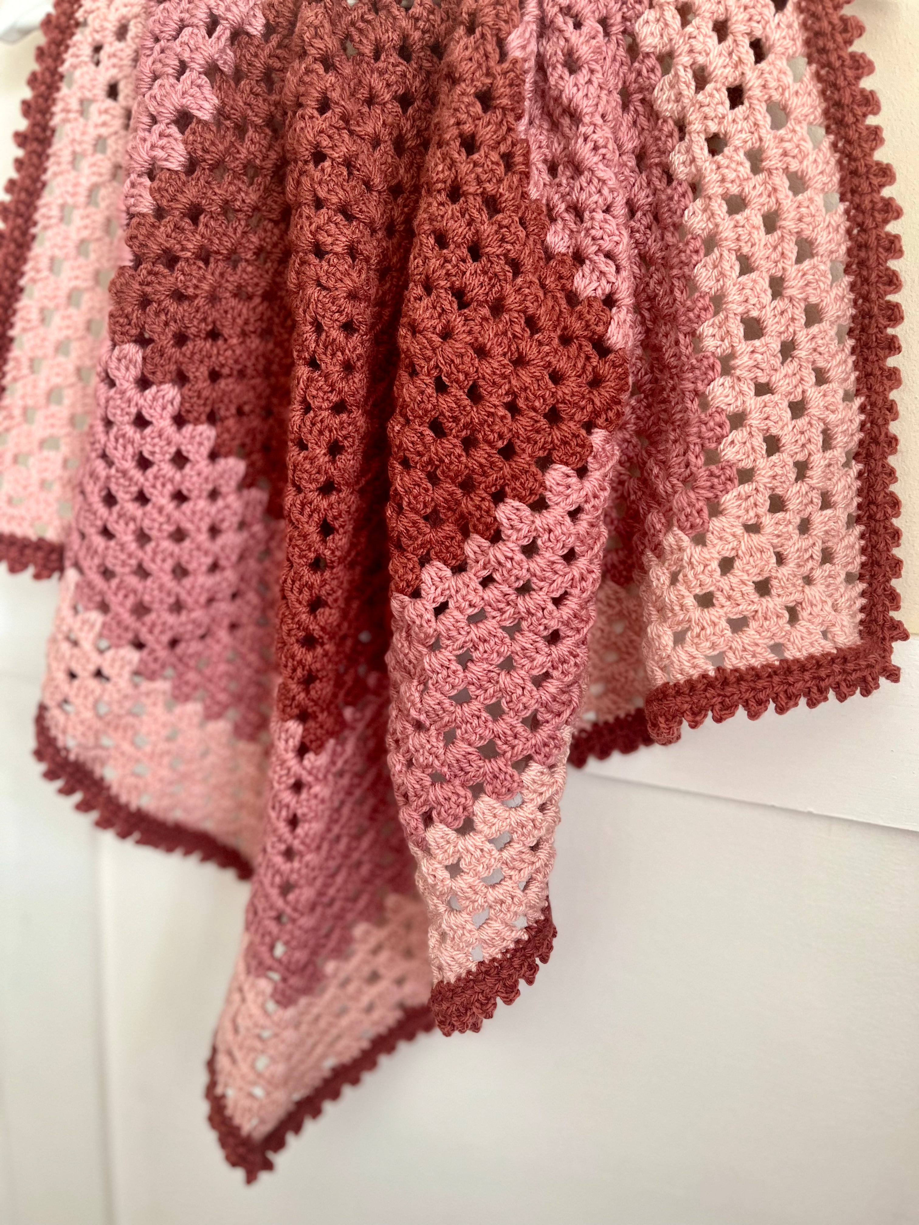 A handmade crocheted blanket draped over a white surface, featuring a patchwork design in shades of dusty rose, terracotta, and light pink with a matching crocheted border.