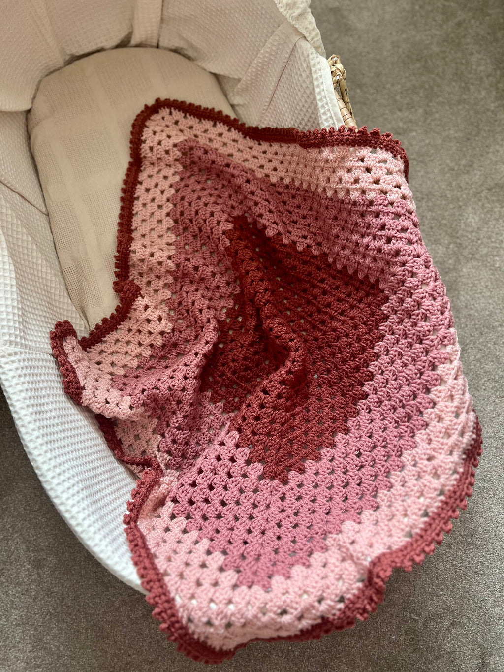A handmade crocheted baby blanket with a gradient color scheme from deep burgundy to light pink, draped over a white bassinet on a gray carpet.