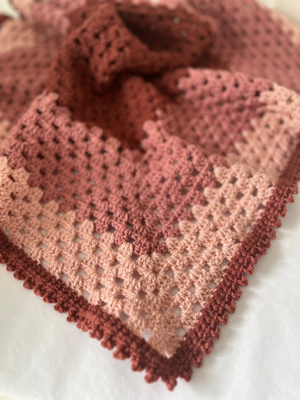 A close-up of a crocheted shawl or scarf with a textured openwork pattern and scalloped edges, shown in a soft gradient of dusty rose, mauve, and terracotta on a white background.