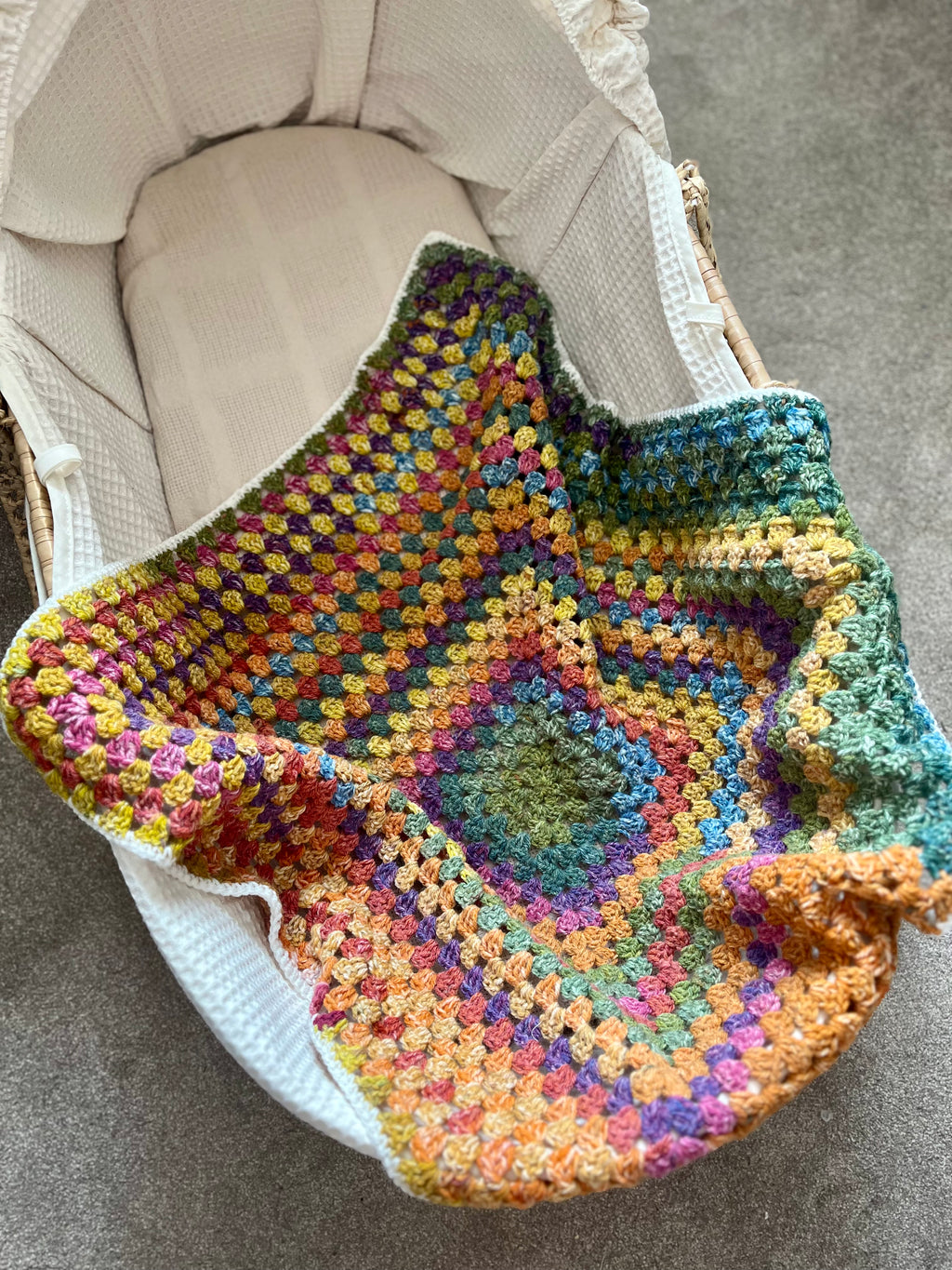 A handmade, multicolored crocheted baby blanket with a starburst pattern, draped over the side of a cream-colored Moses basket on a gray carpet.