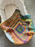 A handmade, multicolored crocheted baby blanket with a starburst pattern, draped over the side of a cream-colored Moses basket on a gray carpet.