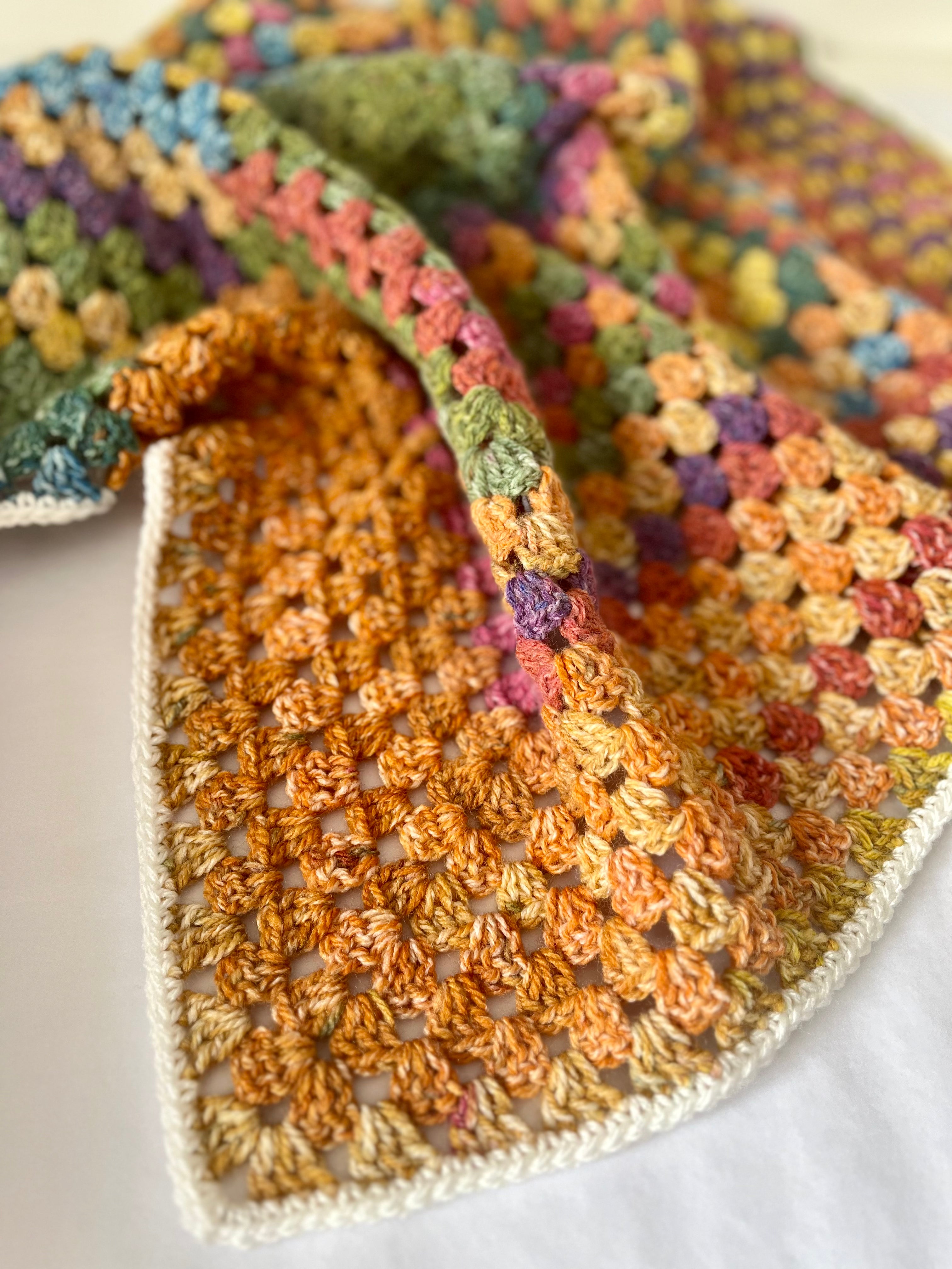 A close-up of a handmade crocheted textile, likely a baby blanket or scarf, featuring a multicolor granny square pattern with a prominent orange section and a white border on a white fabric background.