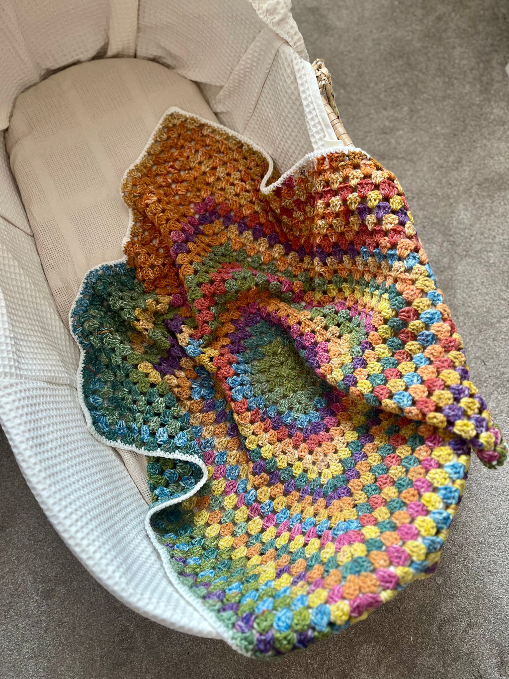 A handmade, multicolored crocheted baby blanket with a rainbow gradient pattern draped over a white woven bassinet.