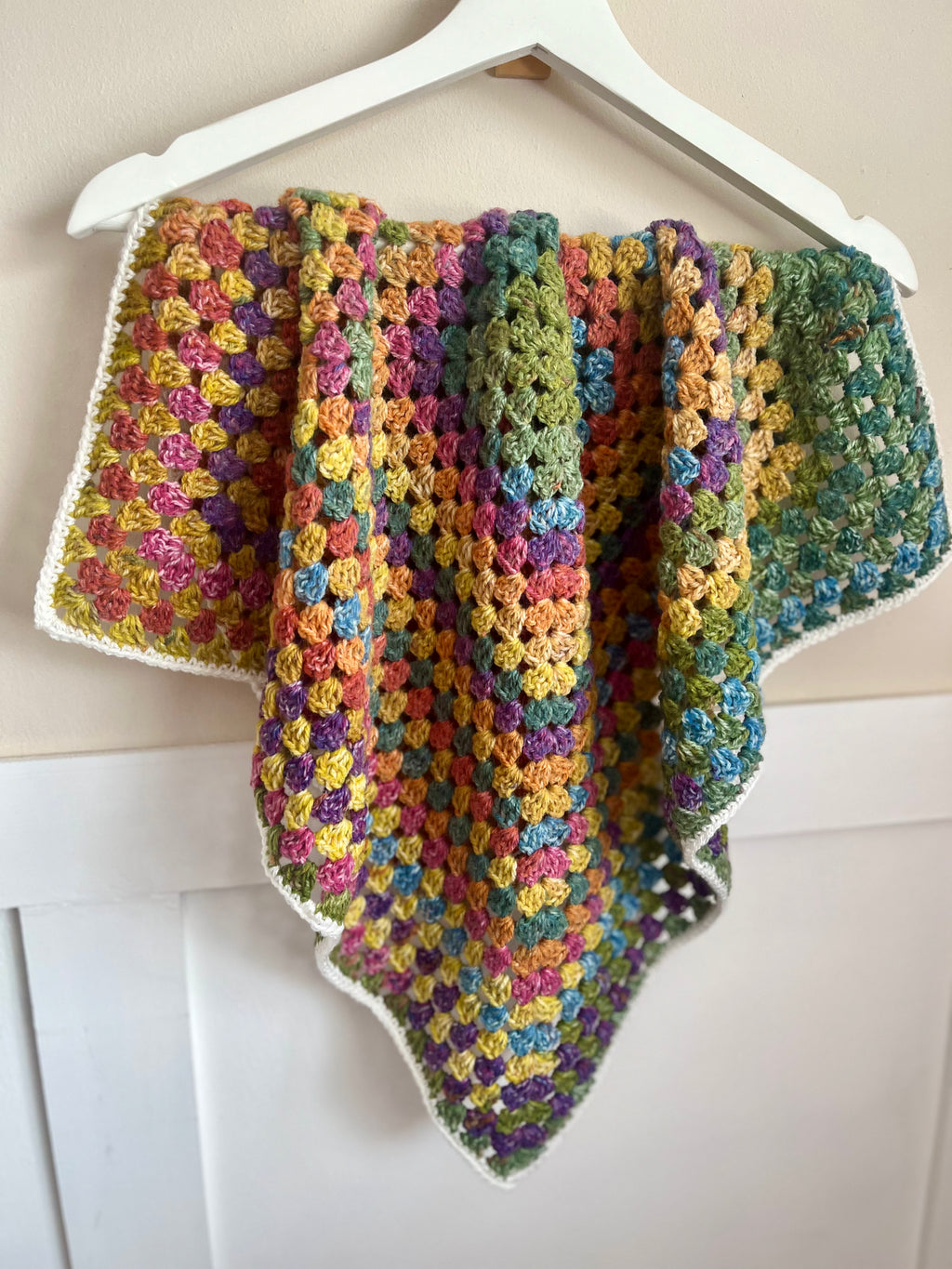 A handmade crocheted baby blanket with a multicolor granny square pattern and a white border, hanging on a white wooden hanger against a light beige wall.