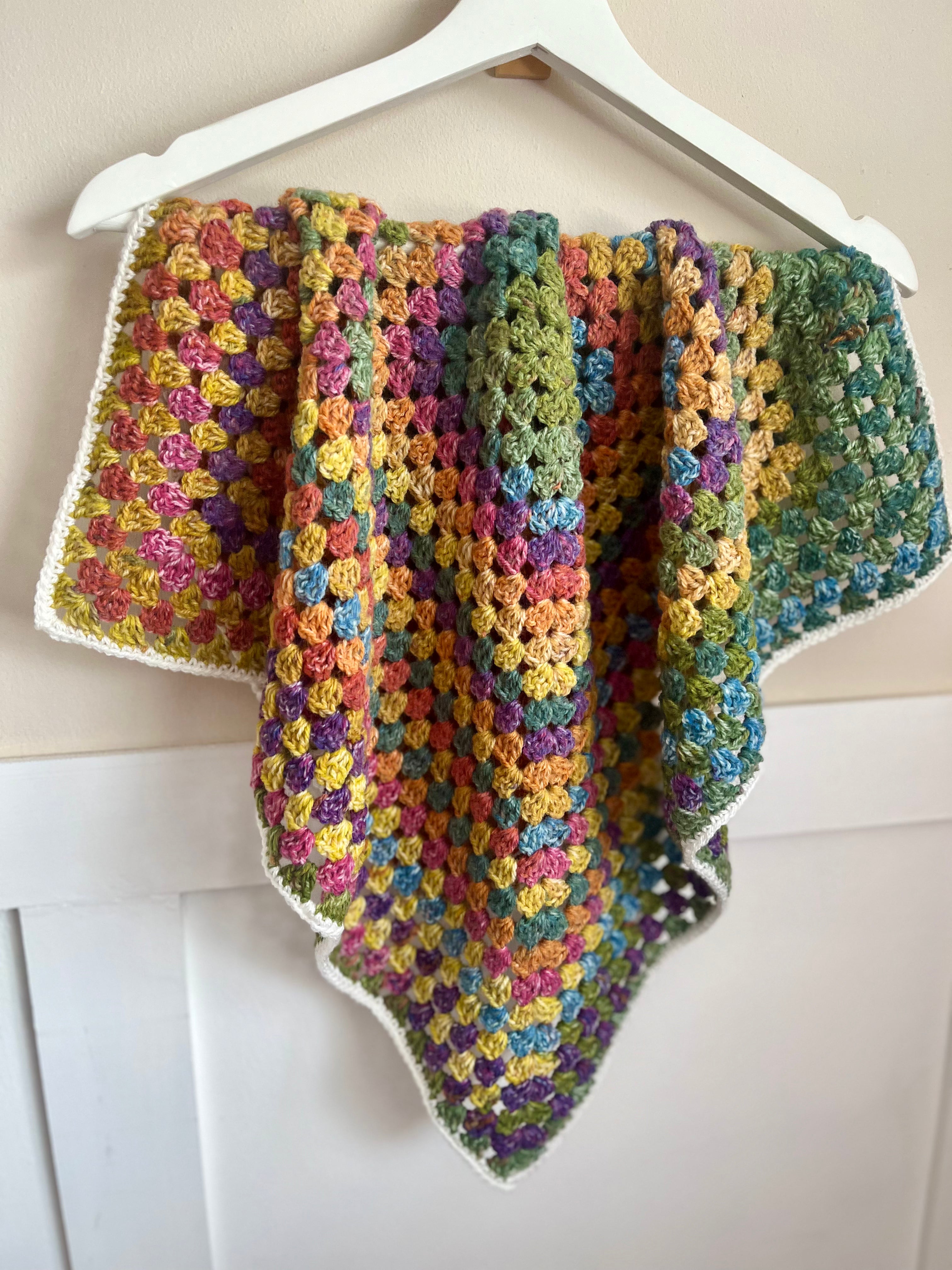 A handmade crocheted baby blanket with a multicolor granny square pattern and a white border, hanging on a white wooden hanger against a light beige wall.