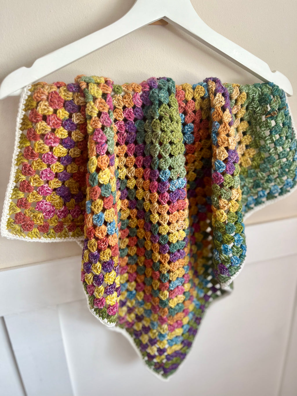 A handmade crocheted shawl or baby wrap with a multicolor rainbow pattern, hanging from a white plastic hanger against a light beige wall.