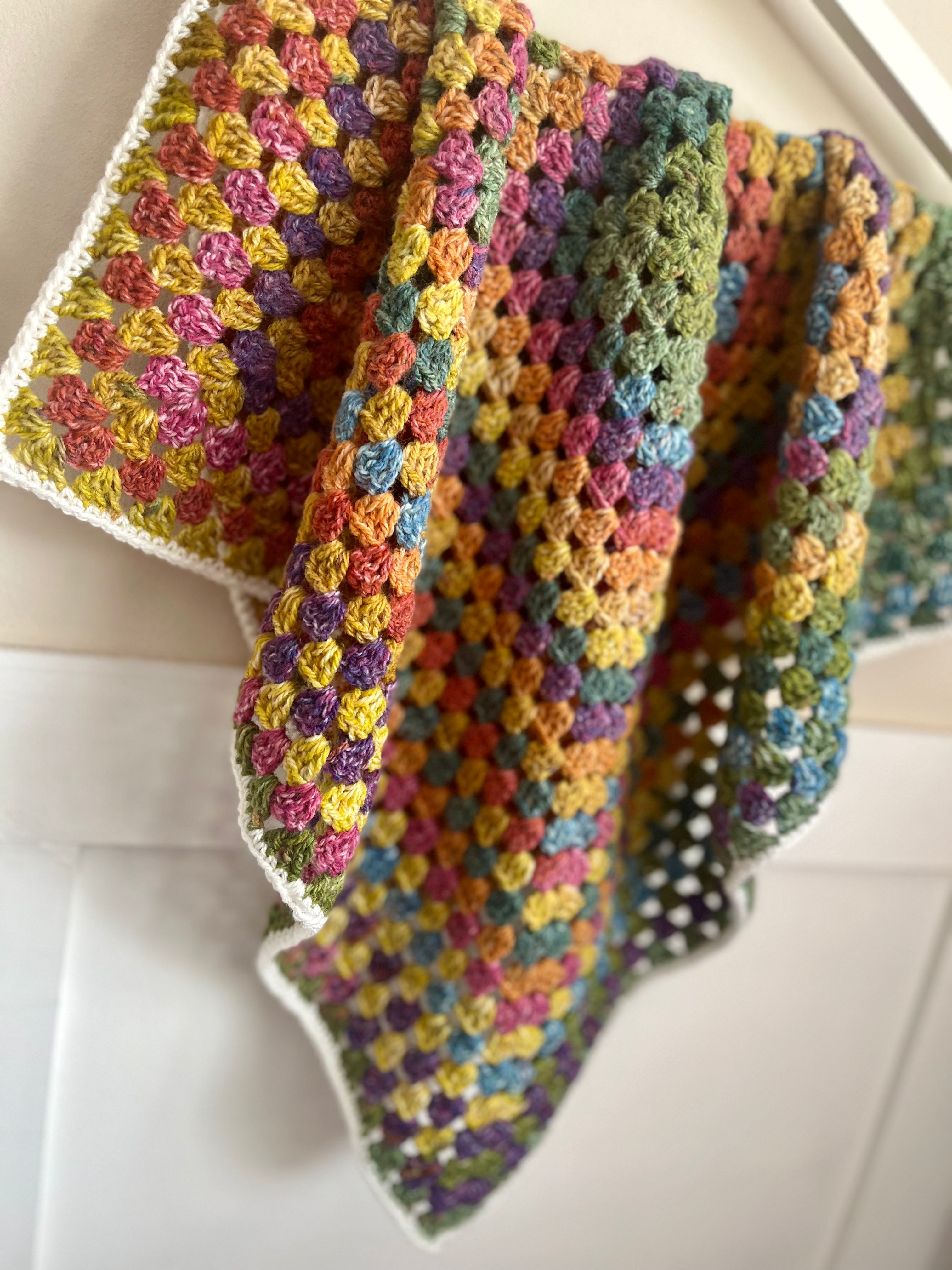 A handmade, multicolored crocheted baby blanket with a white lace border, draped over a white crib or wall.