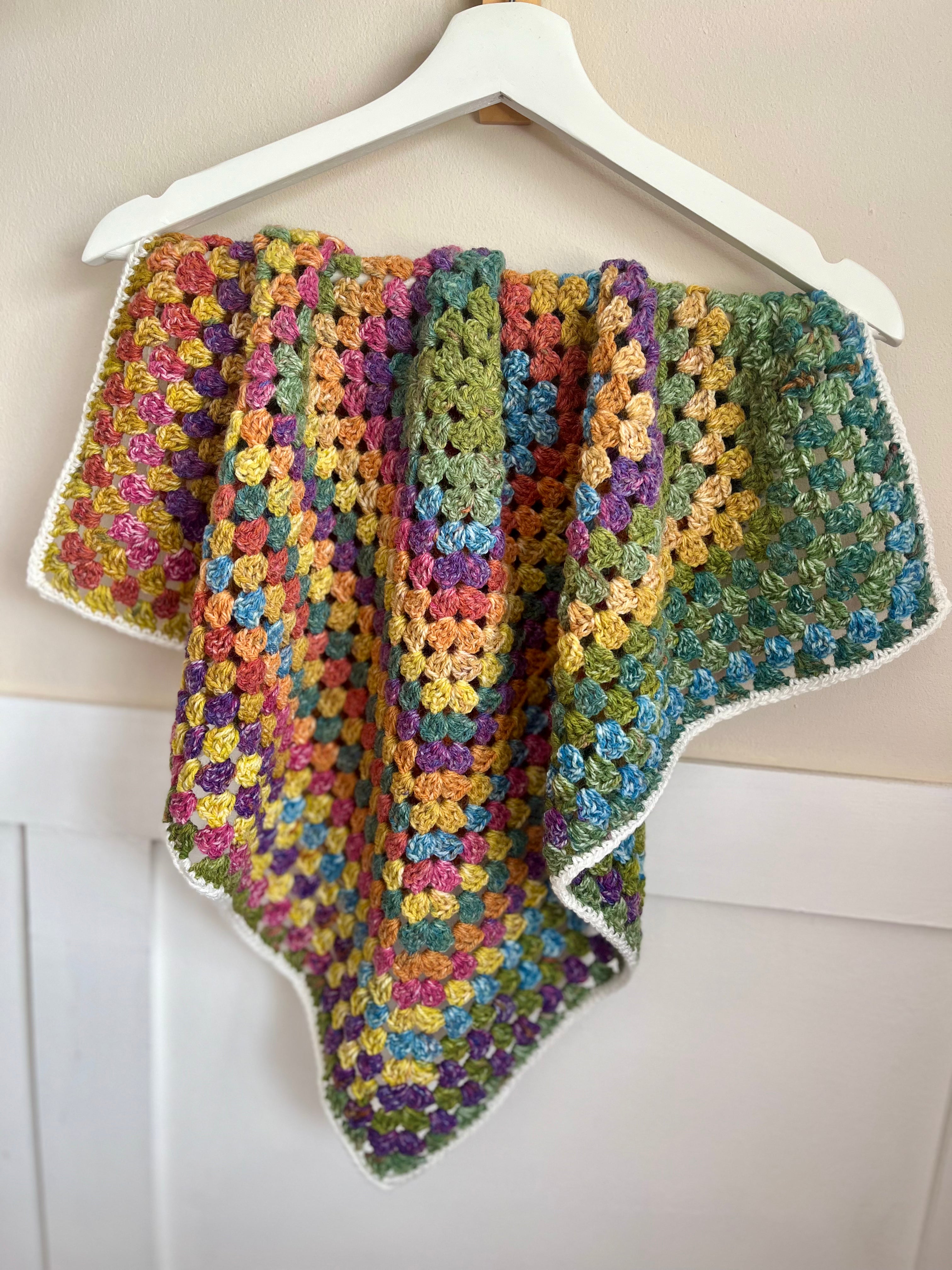 A handmade crocheted baby blanket with a multicolor striped pattern, hanging from a white wooden hanger against a light beige wall.