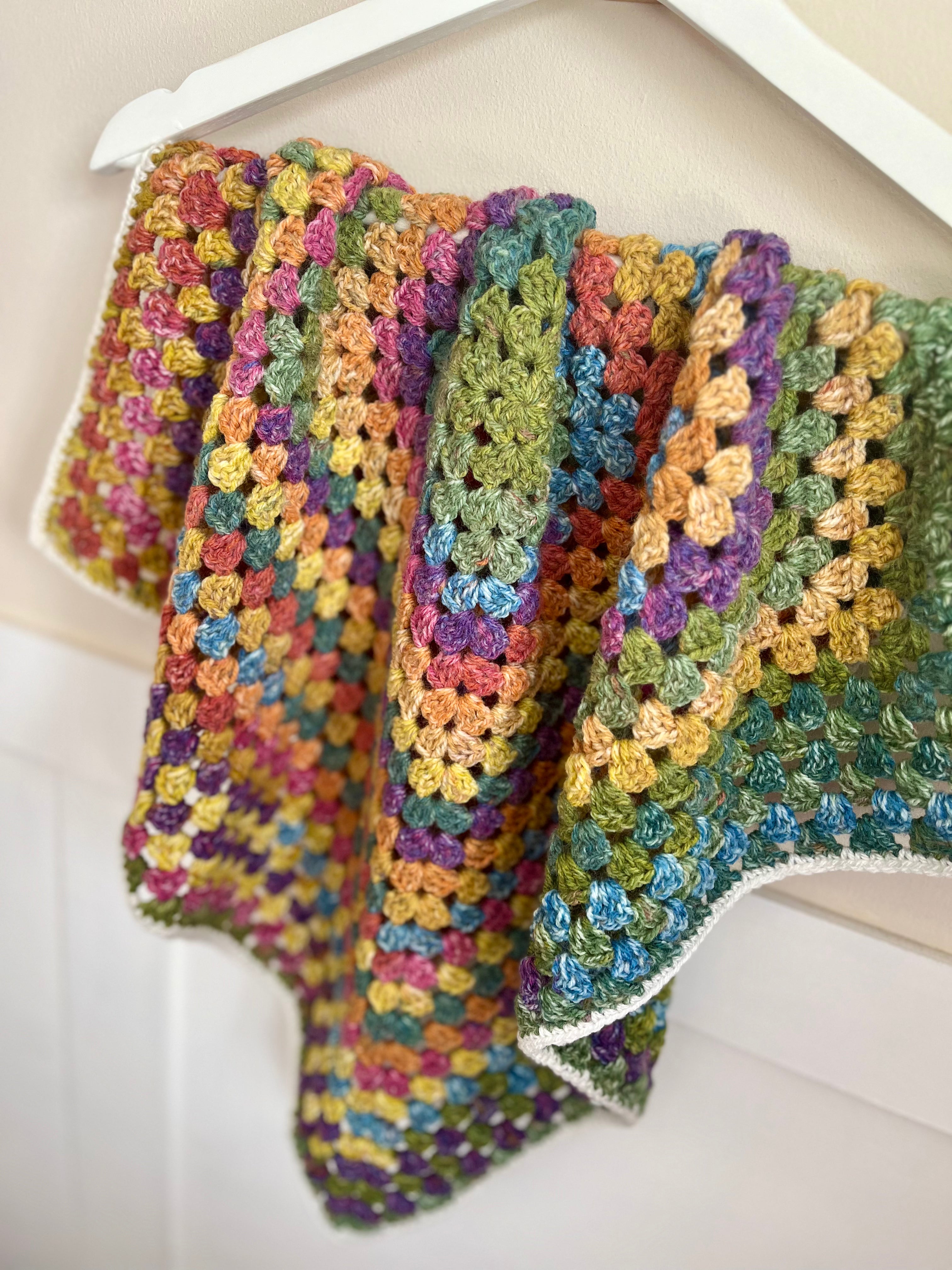 A handmade multicolor crochet blanket draped over a white hanger against a light beige wall.