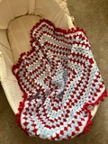 A handmade crocheted blanket with red, white, and light blue stripes and pom-pom trim, draped over a beige bassinet.