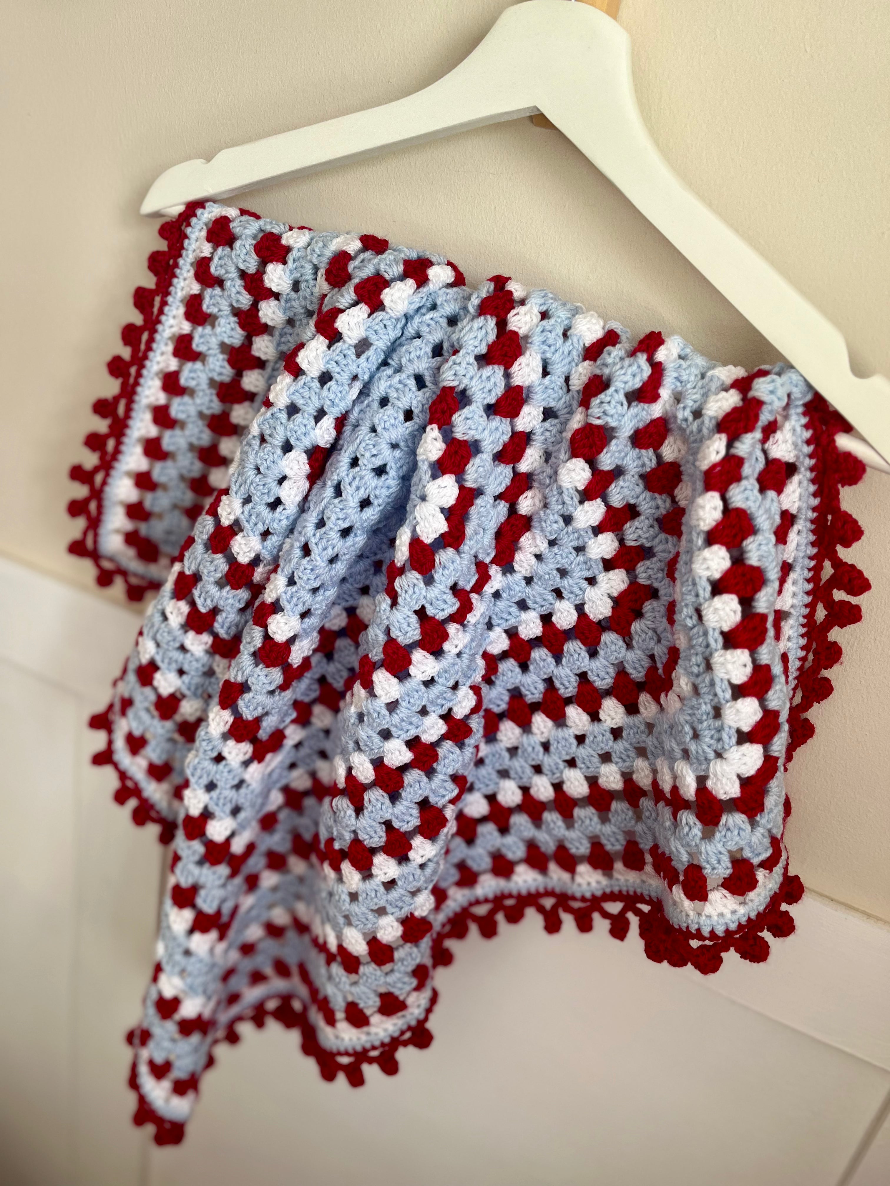 A crocheted baby blanket with a red, white, and light blue granny square pattern and pom-pom trim, draped over a white plastic hanger against a cream-colored wall.
