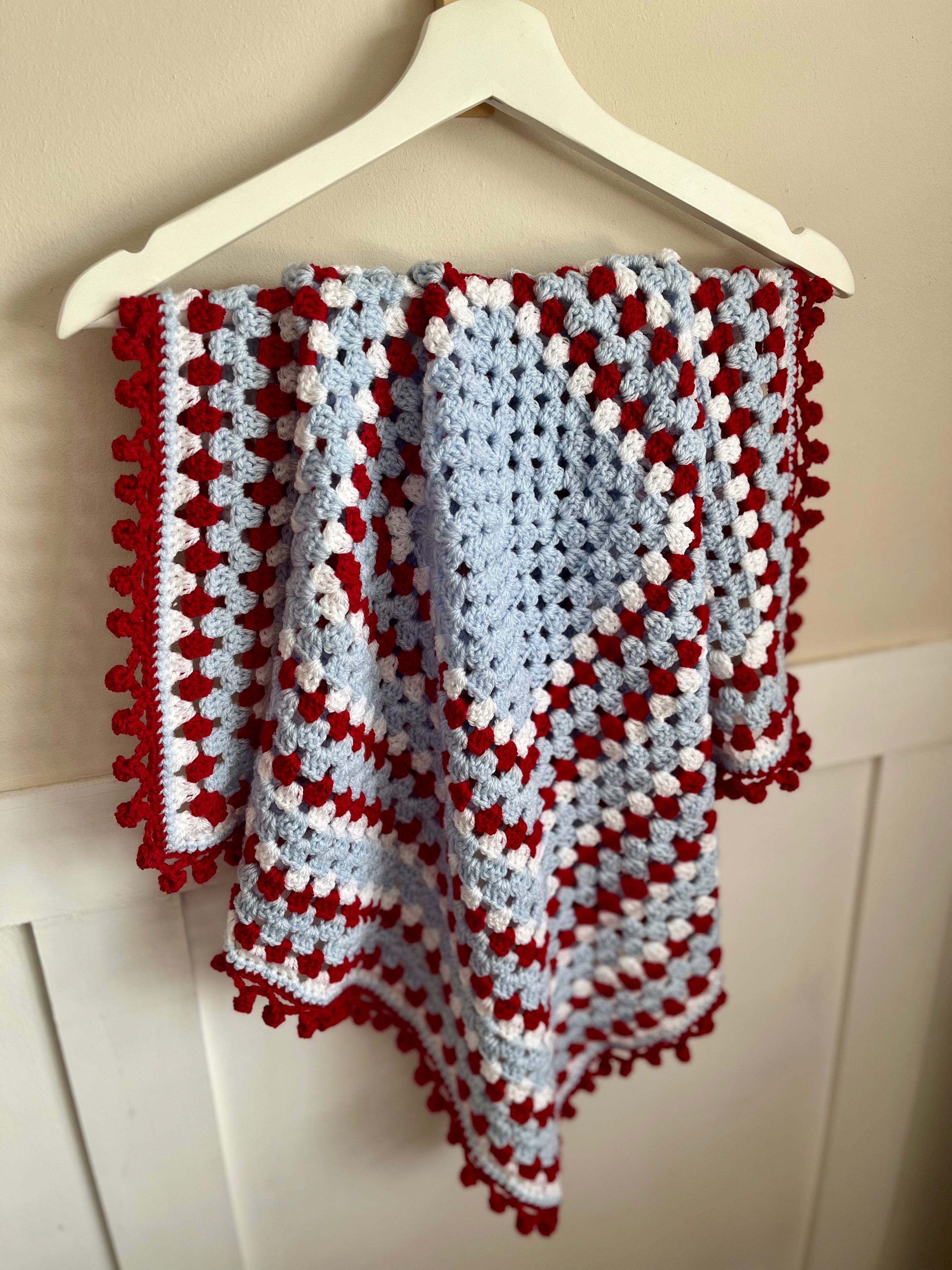 A handmade crocheted baby blanket with a red and white pom-pom border and a central light blue heart motif, hanging on a white wooden hanger against a cream wall.