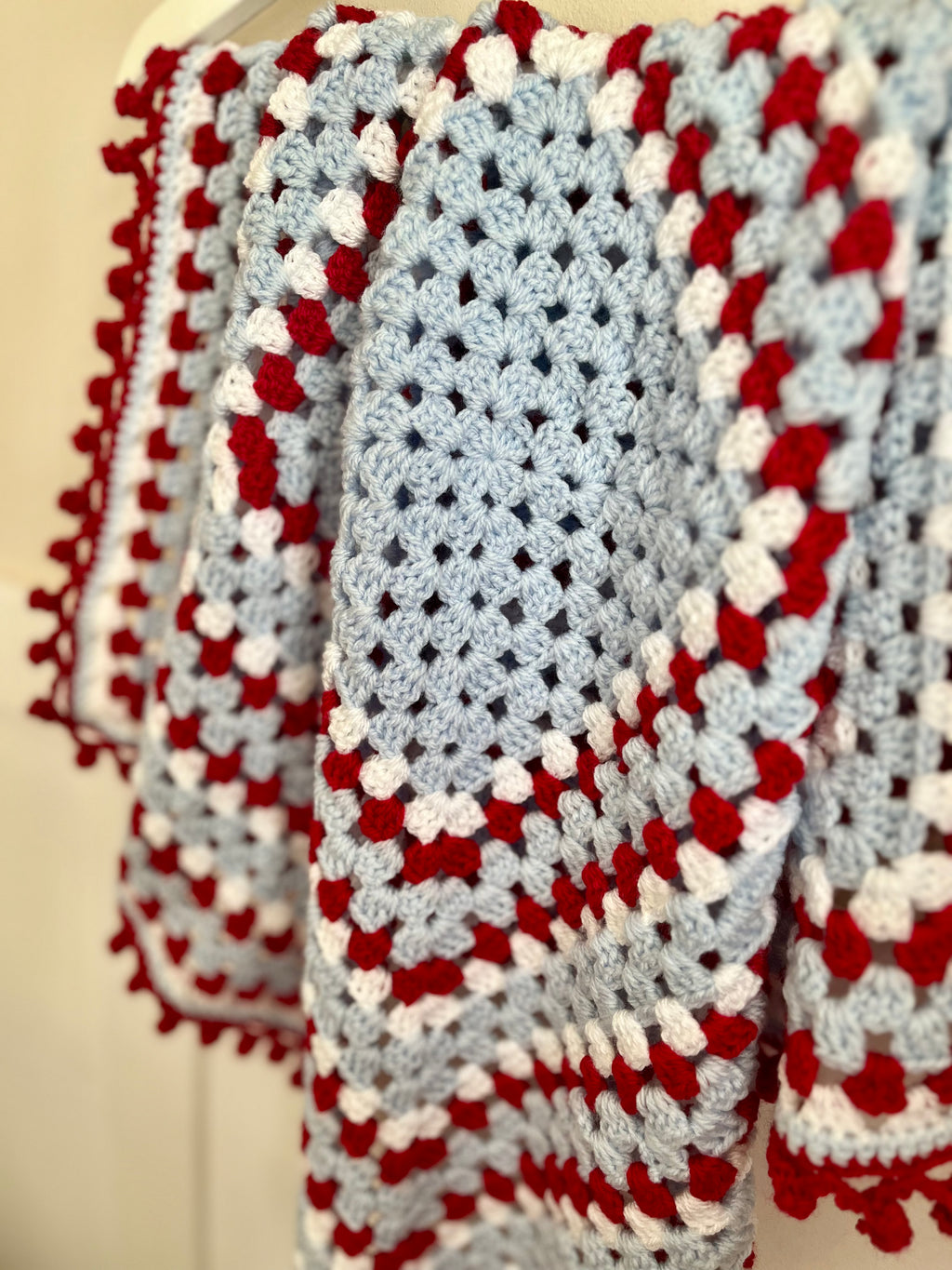 A close-up of a handmade crocheted blanket draped over a hanger, featuring a light blue center with red and white textured stripes and pom-pom trim.