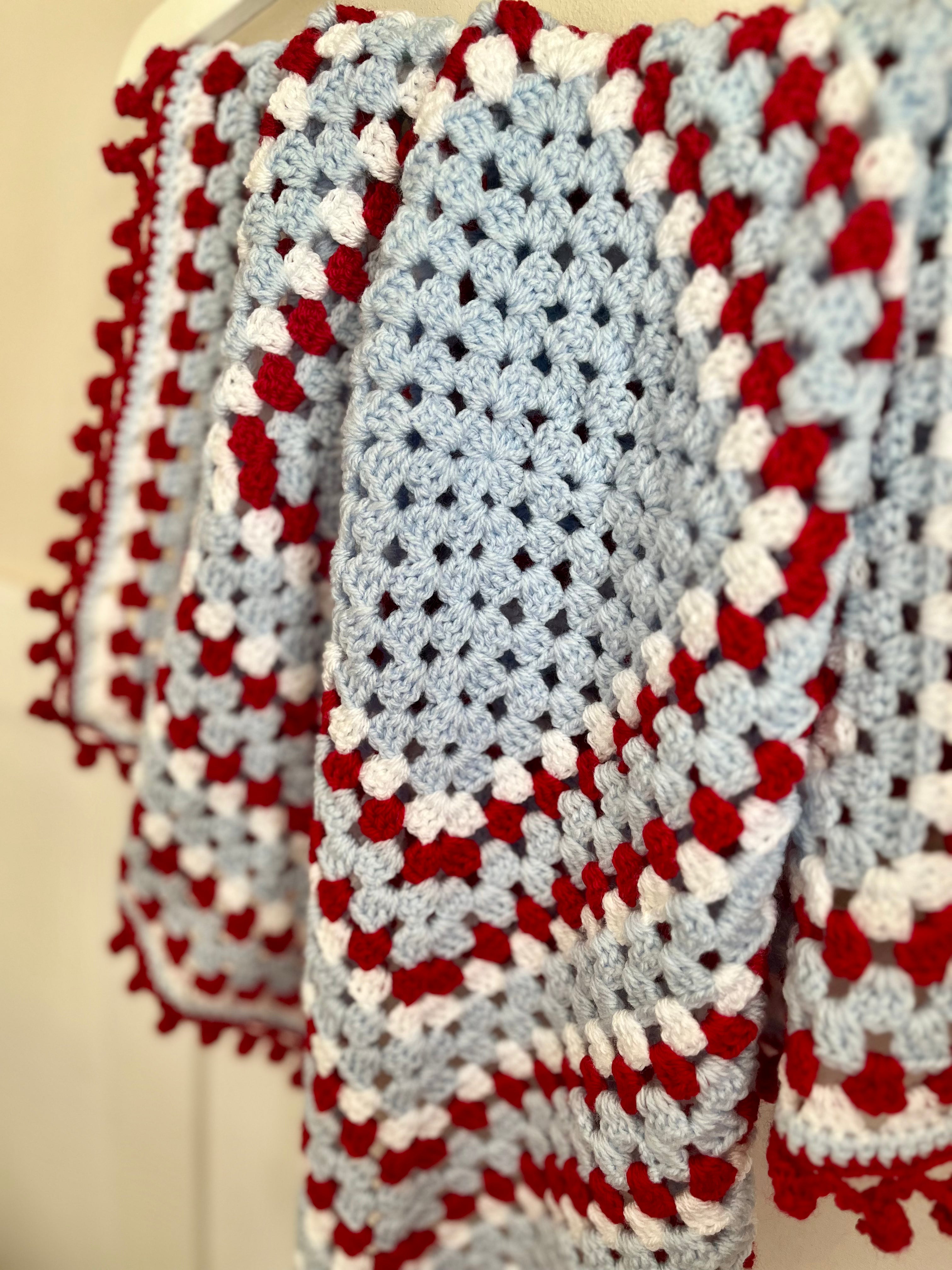 A close-up of a handmade crocheted blanket draped over a hanger, featuring a light blue center with red and white textured stripes and pom-pom trim.
