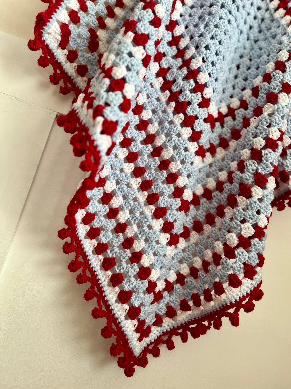 A handmade crocheted baby blanket with a red pom-pom border, featuring a pattern of light blue, white, and red granny squares on a cream background.