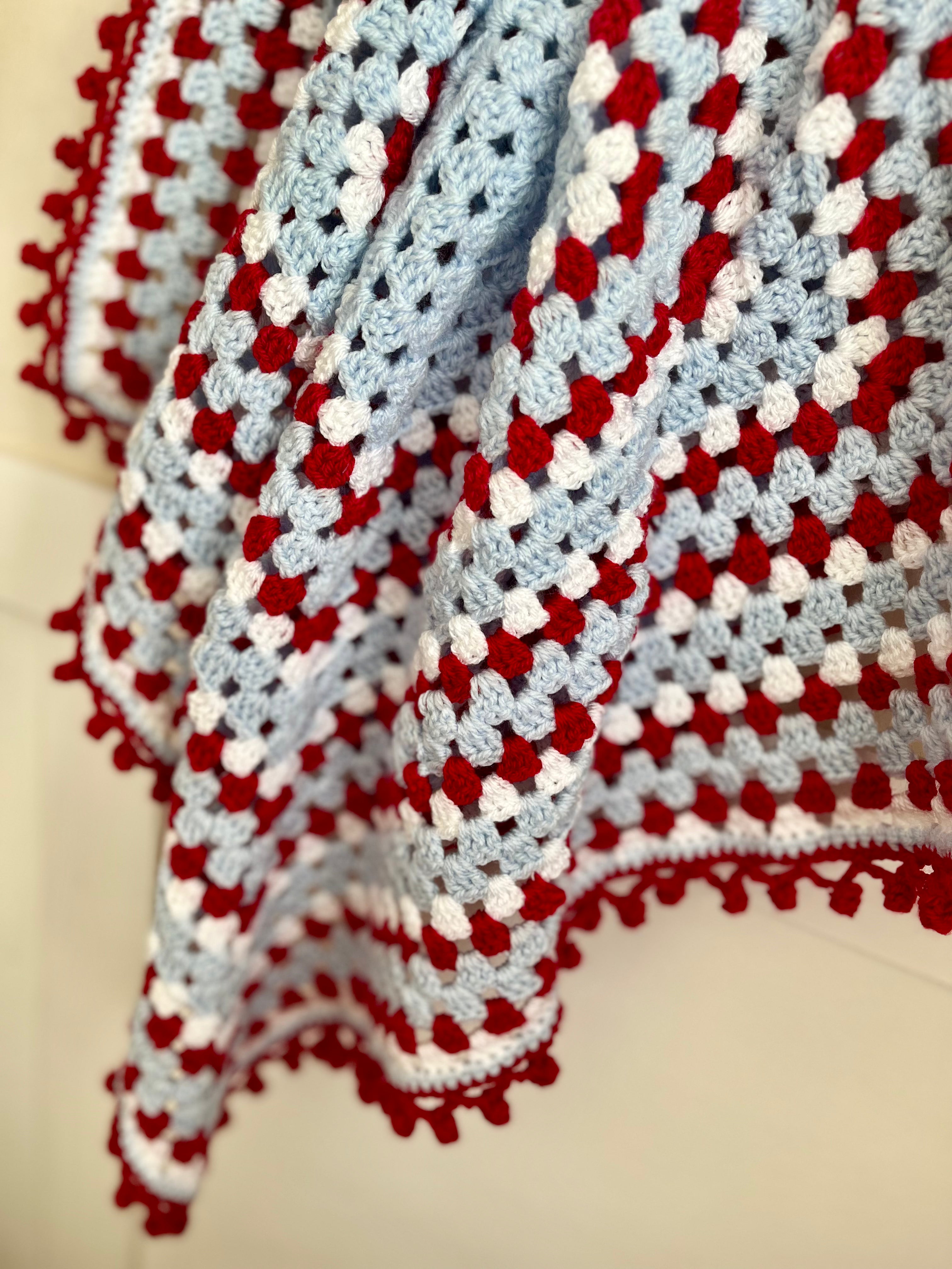 A close-up of a handmade crocheted blanket featuring a textured pattern of red, light blue, and white yarn with a pom-pom trim.