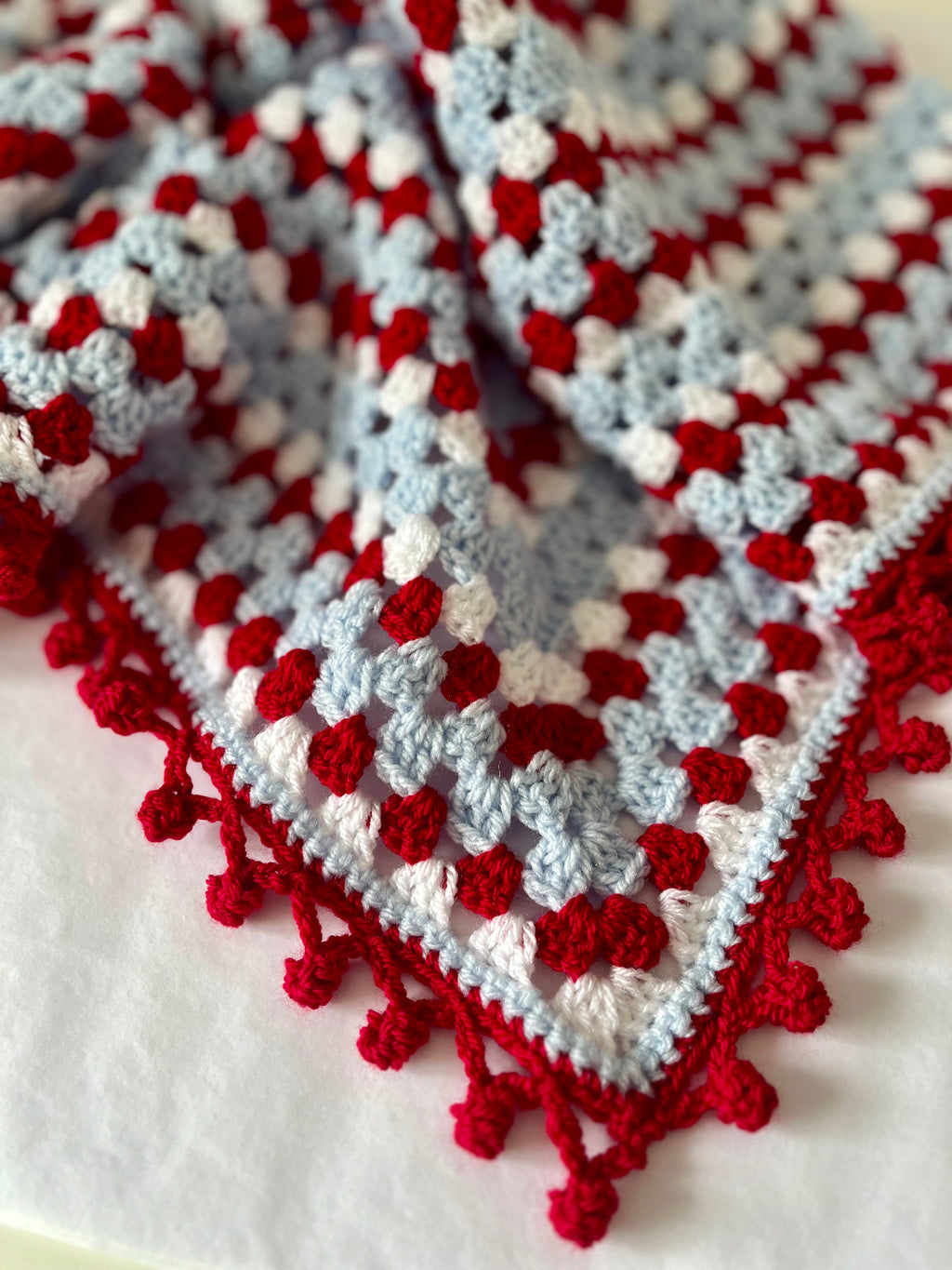 A close-up of a crocheted textile with a granny-square pattern in light blue, white, and red, finished with a red pom-pom border, laid on a white surface.