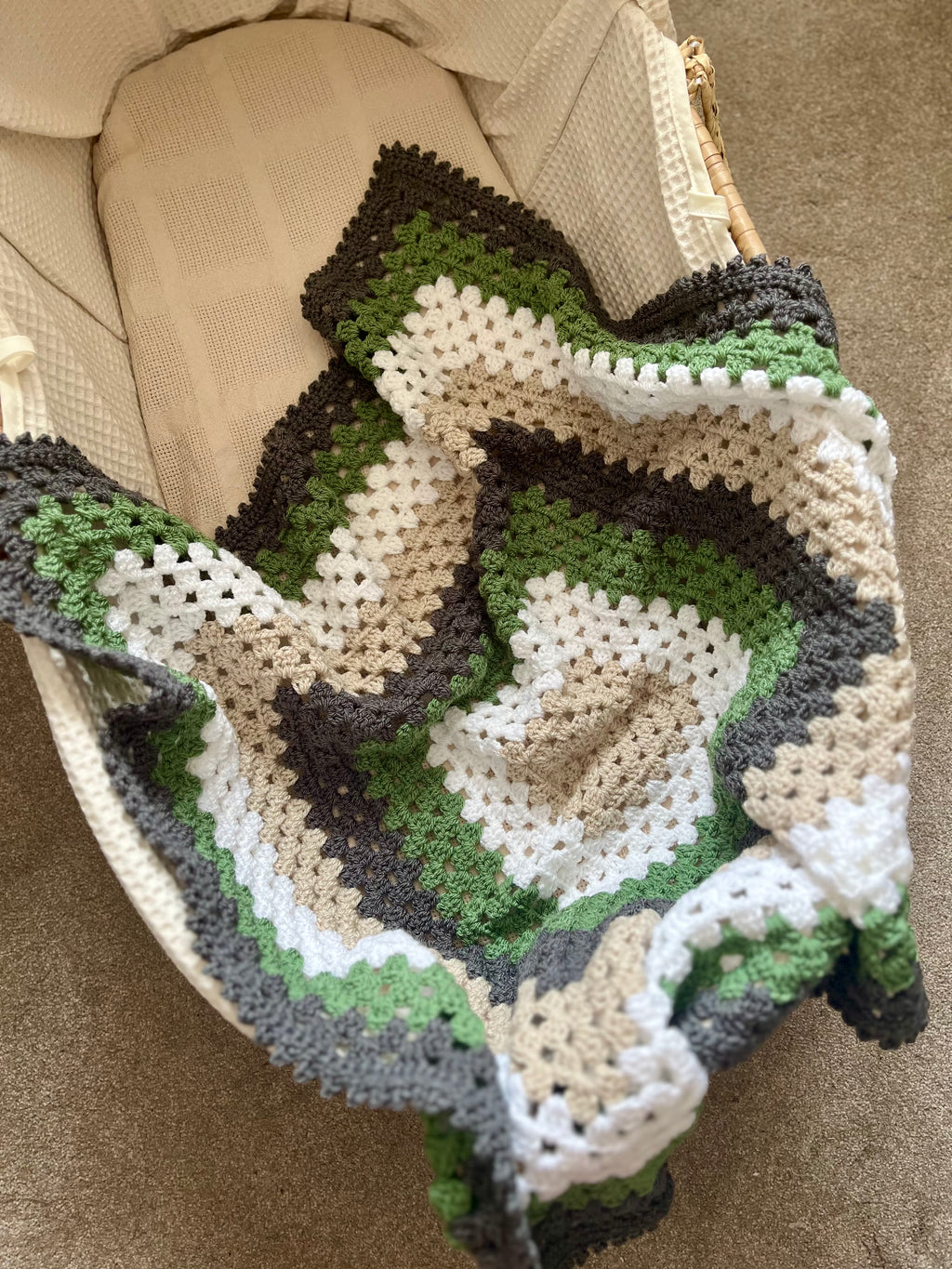 A handmade crocheted baby blanket with concentric rings of green, beige, white, and dark brown, draped over a cream-colored bassinet.