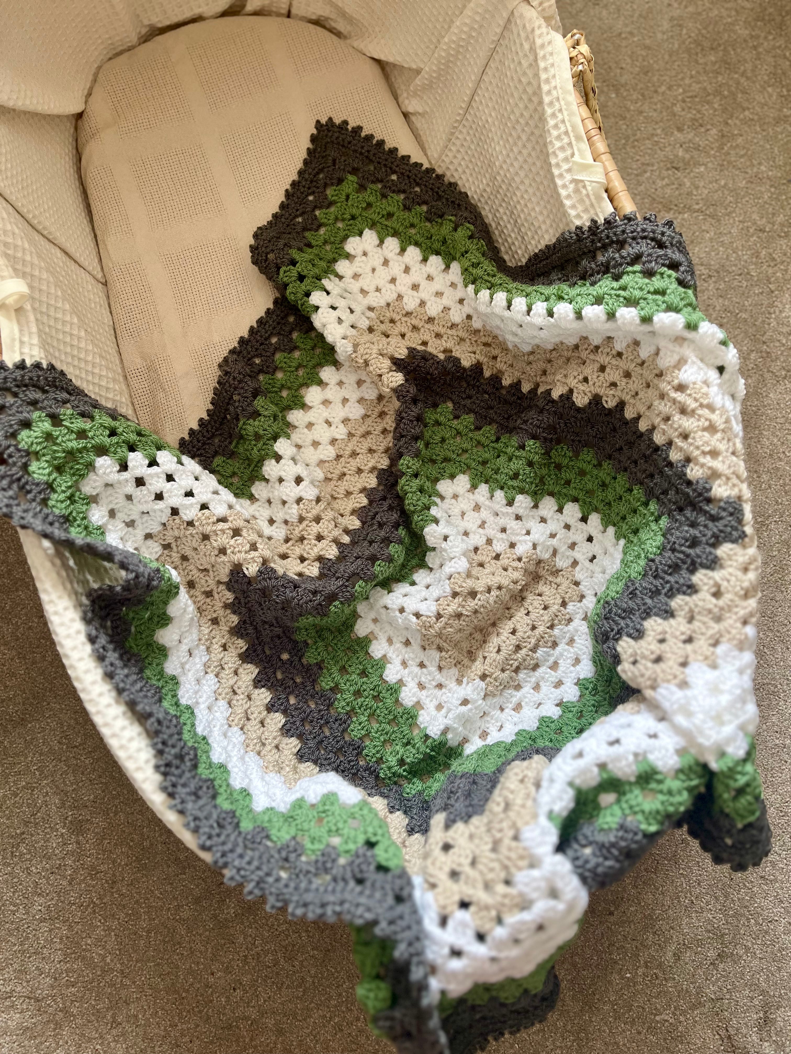 A handmade crocheted baby blanket with concentric rings of green, beige, white, and dark brown, draped over a cream-colored bassinet.