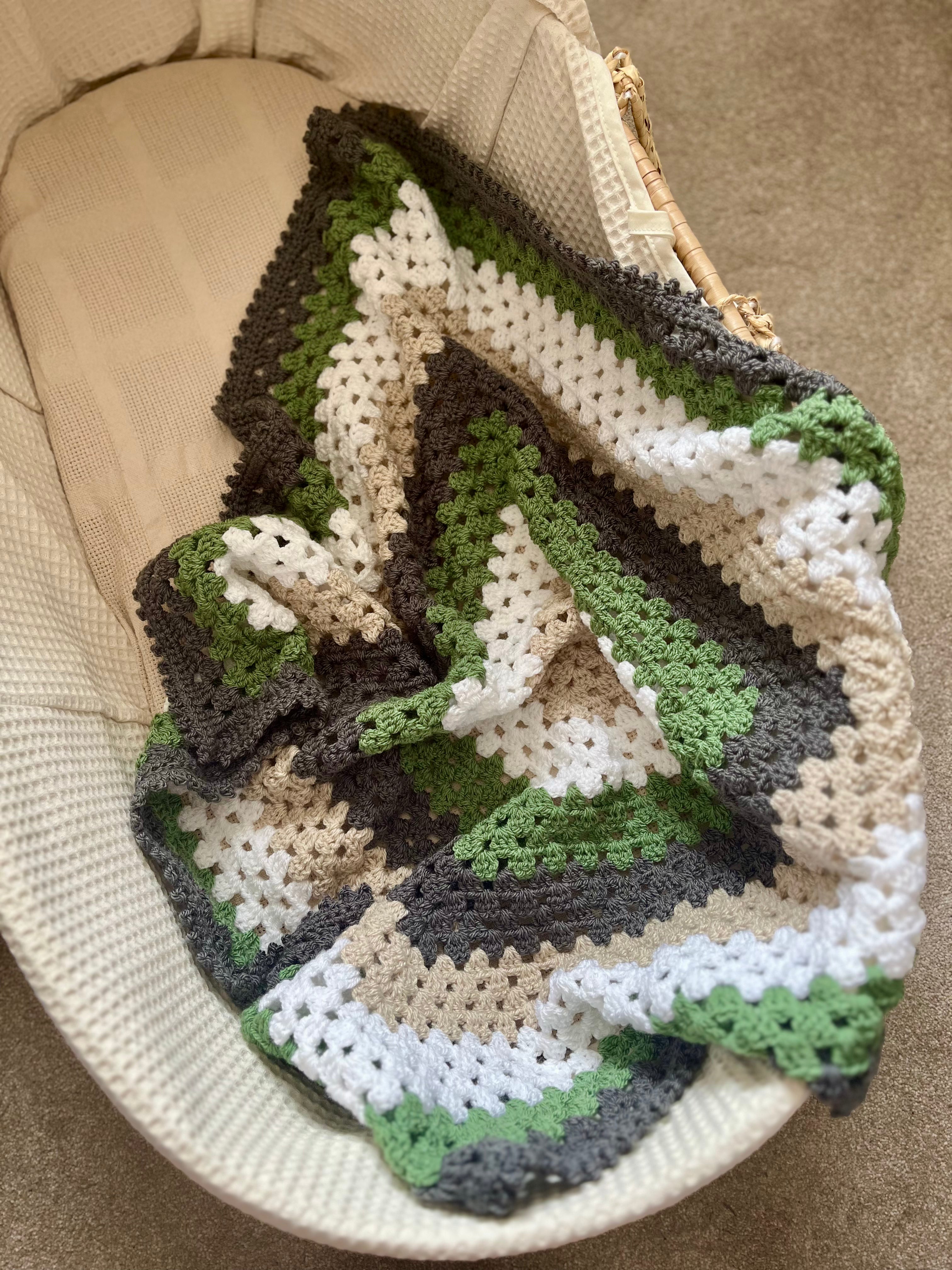 A handmade crocheted baby blanket with a green, brown, beige, and white geometric pattern, draped over a cream-colored woven bassinet.