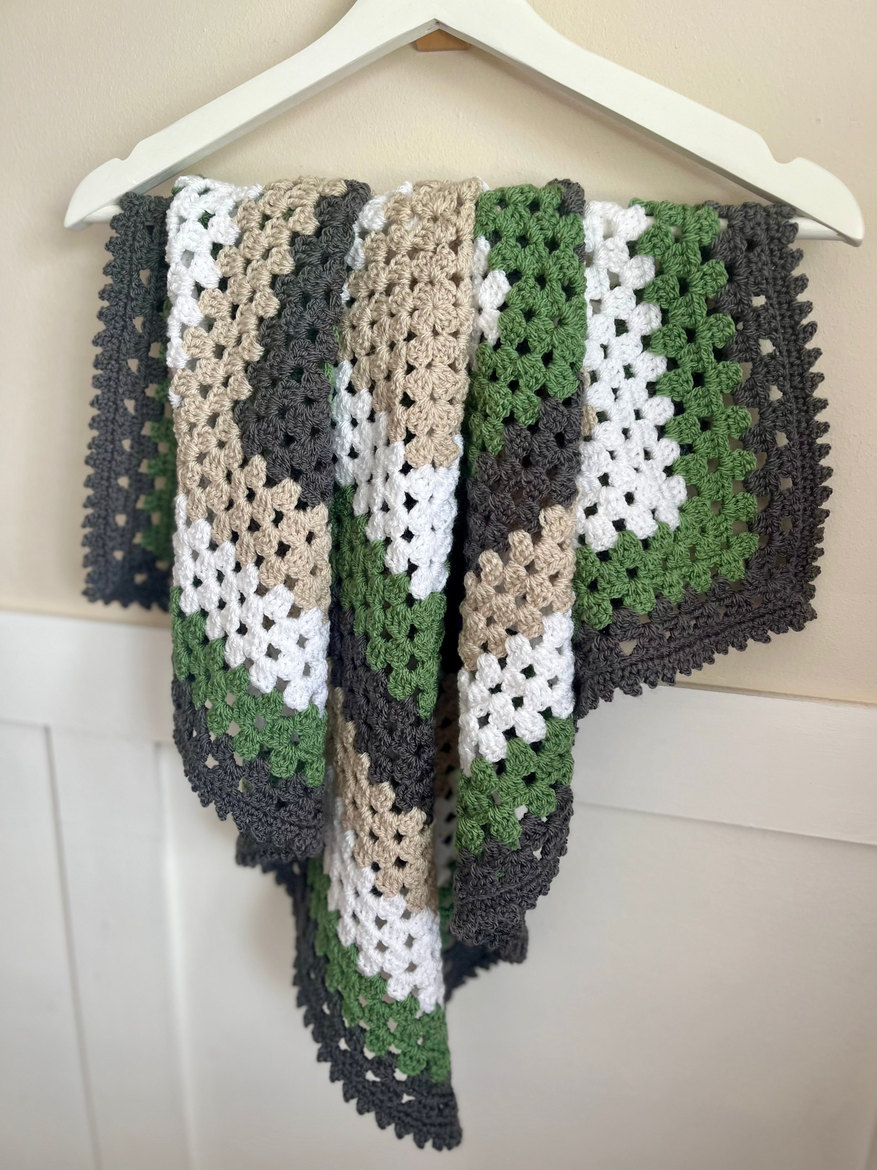 A handmade crocheted shawl or blanket with a geometric pattern in green, dark gray, beige, and white, draped over a white wooden hanger against a light beige paneled wall.