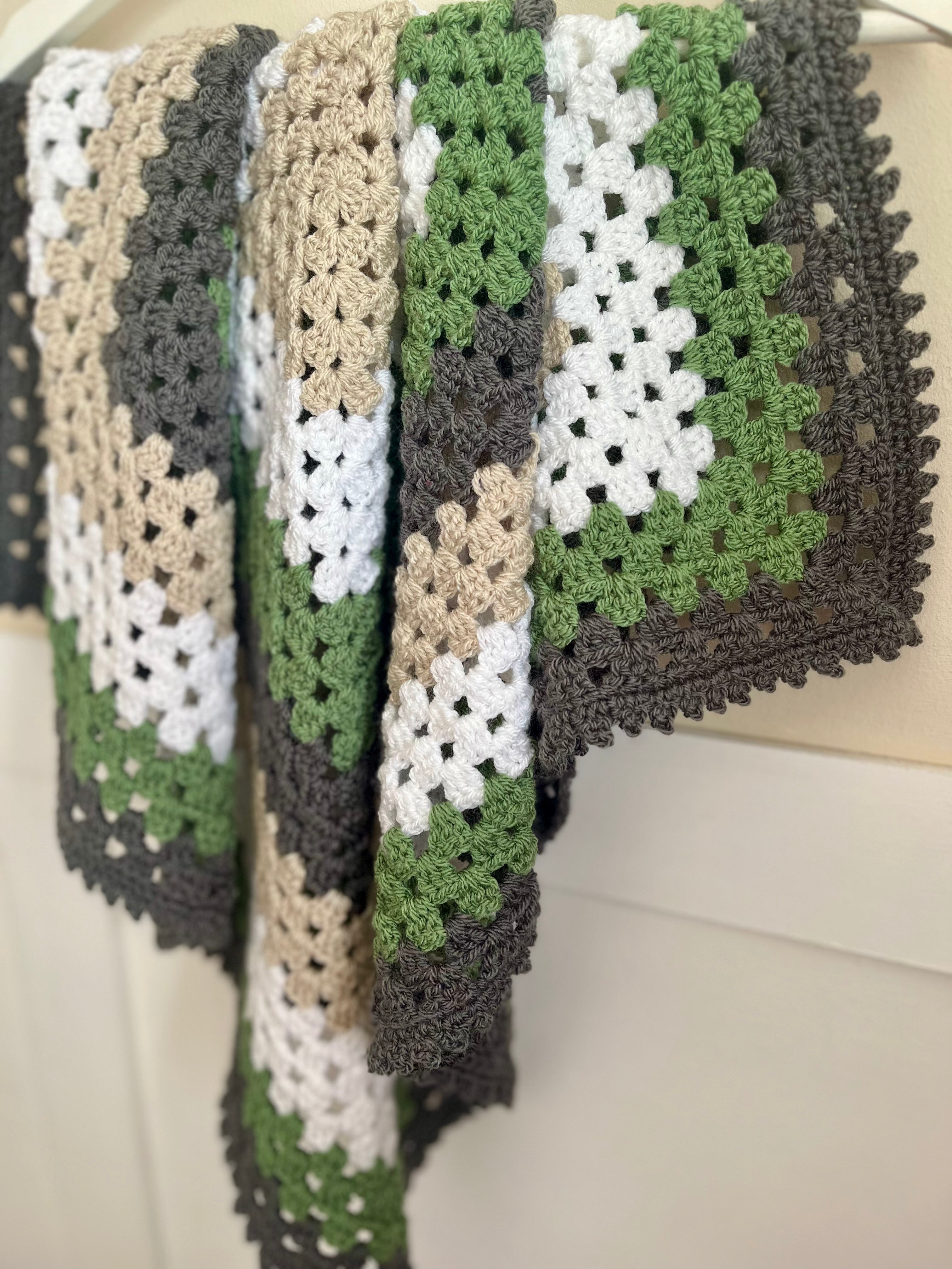 A handmade crocheted granny square blanket draped over a white surface, featuring a geometric pattern of green, white, beige, and dark gray yarn.
