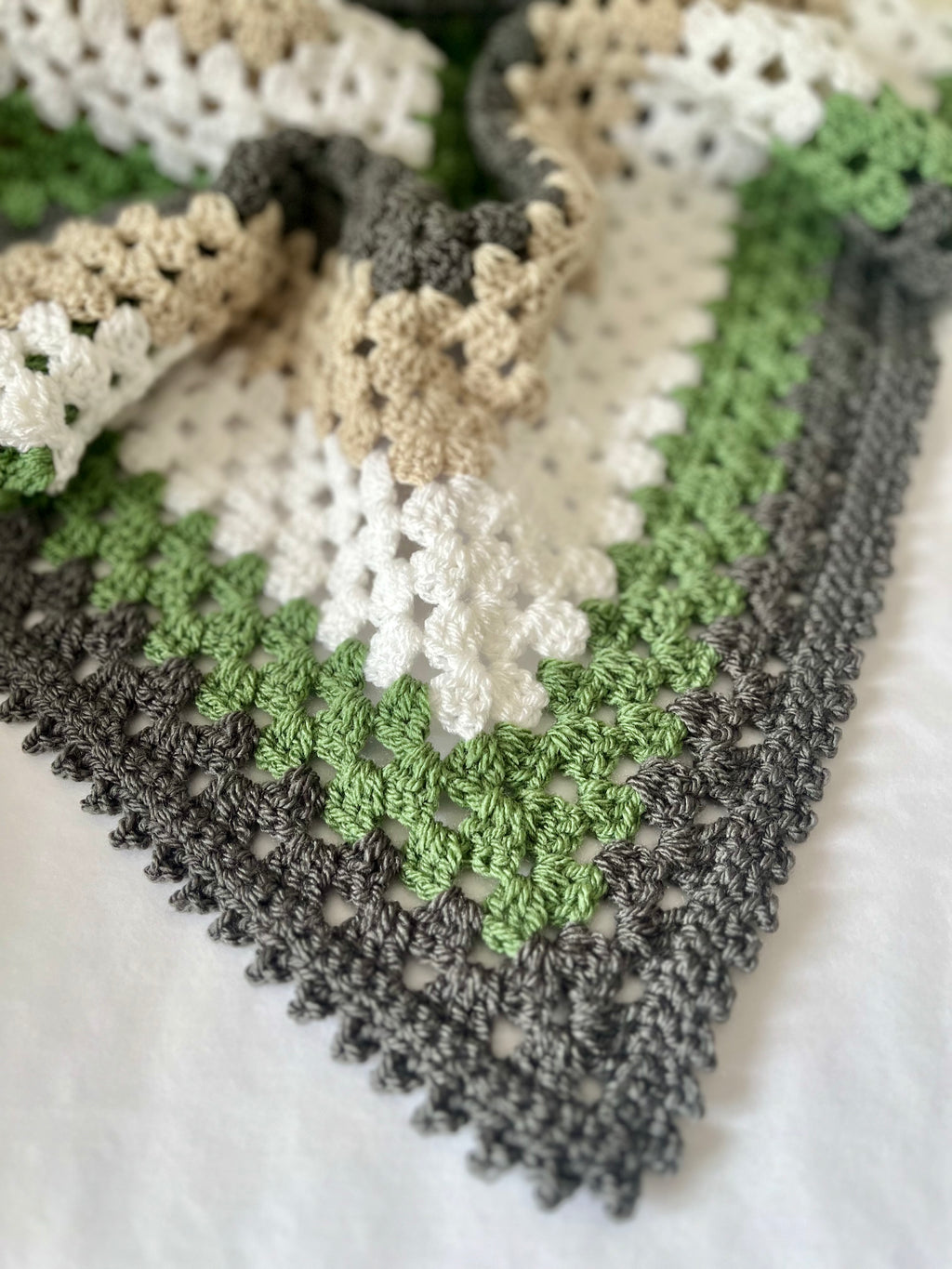 A close-up of a crocheted blanket with a striped pattern in dark gray, light green, and off-white, laid on a white fabric background.
